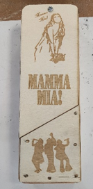 Photograph of a wooden plaque featuring engraved designs related to "Mamma Mia!" with a woman holding flowers at the top and three dancing figures at the bottom.