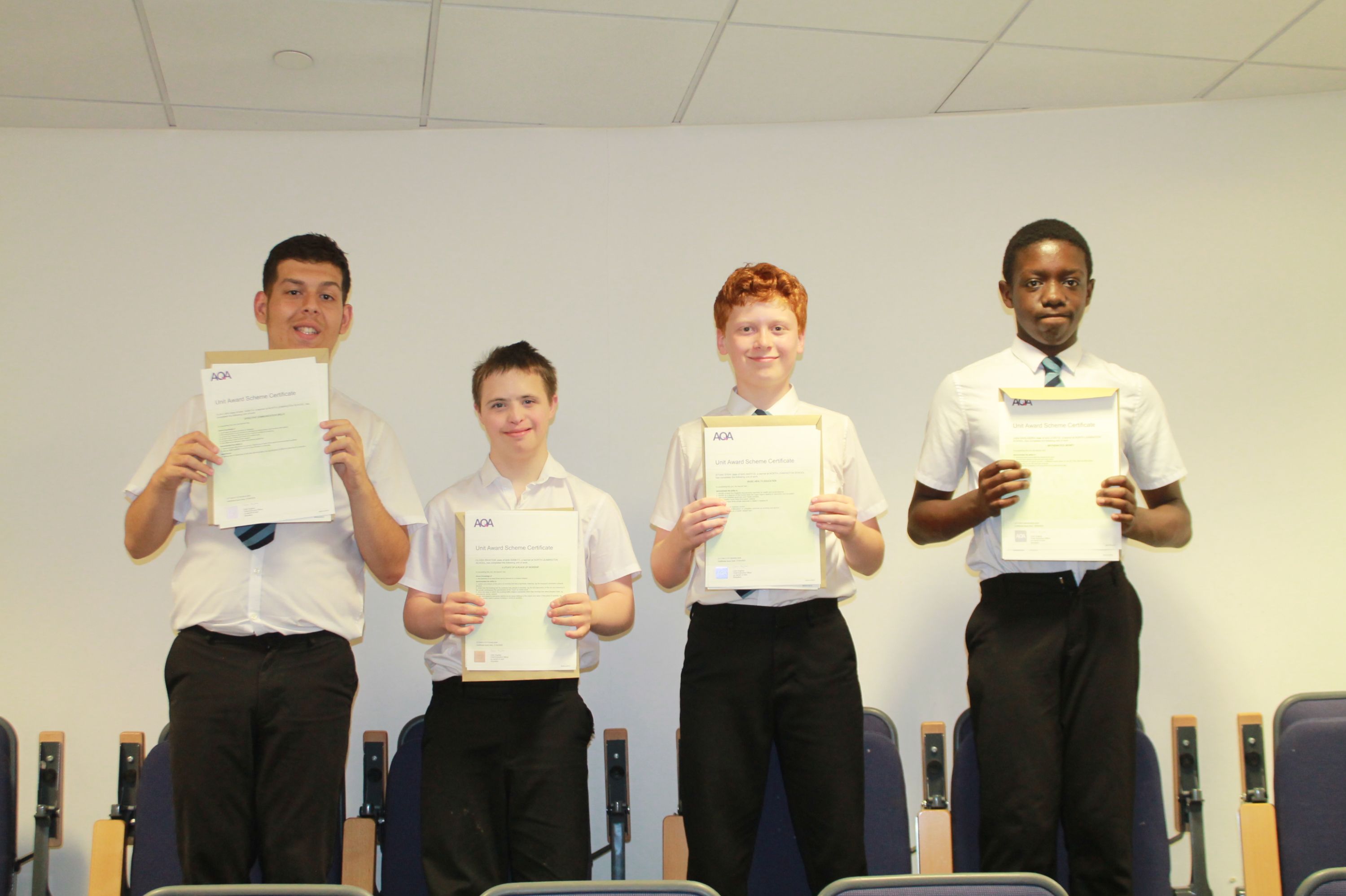 Photograph of four boys standing in a row holding certificates, likely celebrating an academic or extracurricular achievement. They wear white shirts and black trousers, with a plain background and chairs partially visible behind them