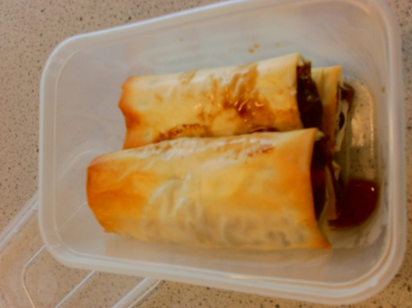  two spring rolls placed inside a transparent plastic container on a speckled countertop. Spring rolls have golden-brown, crispy wrappers with some sauce visible on top and around edges.