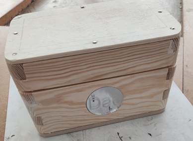 Photograph of a handcrafted wooden box with a hinged lid, featuring visible wood grain and finger joint corners for sturdy construction. A circular cutout on the front reveals part of a label inside, and the box rests on a white surface with screws securing the lid.