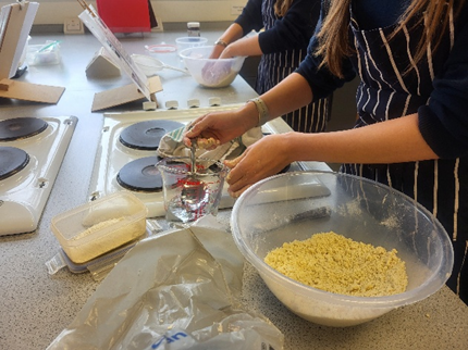 Photograph showing people preparing dough in a kitchen setting with electric stovetops and various baking ingredients on a countertop. Key elements include a large clear bowl filled with yellow dough mixture, a measuring jug with water, and a container of flour, highlighting a baking or cooking activity in progress.