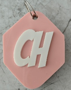 Photograph of a pink, hexagonal keychain with white raised letters "CH" attached to a metal ring. Keychain rests on a white surface