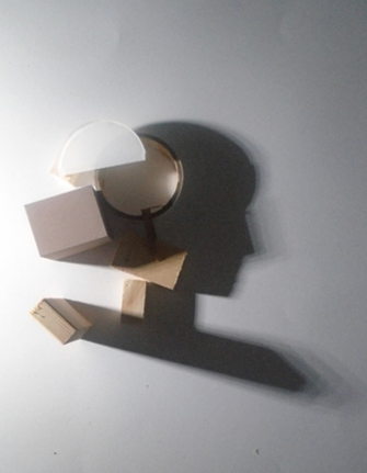 Photograph showing wooden blocks and a white plastic cup arranged on a flat surface, casting distinct shadows. The composition highlights geometric shapes and interplay of light and shadow, with shadows forming elongated patterns extending from the objects.