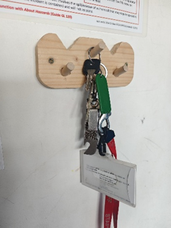 Photograph of a wooden key holder mounted on a white wall, featuring four pegs with multiple keys and keychains hanging from two pegs. A red lanyard with an identification card is attached to one key, and a safety instruction poster is partially visible above the holder