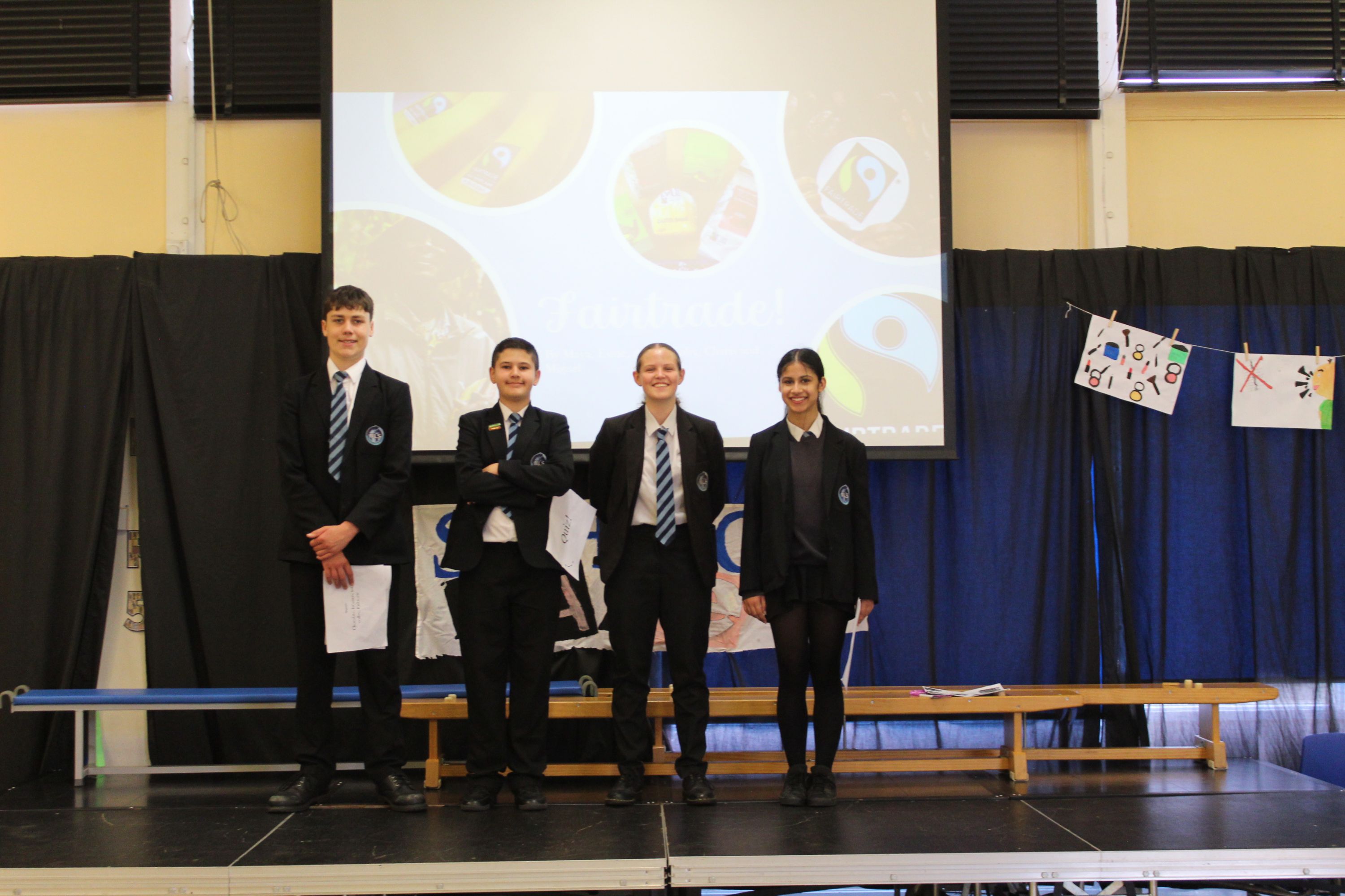 Photograph of four students standing on a stage in school uniforms, likely during a presentation or assembly. Background includes a projection screen displaying circular graphics and a banner with student artwork hanging on a blue curtain.