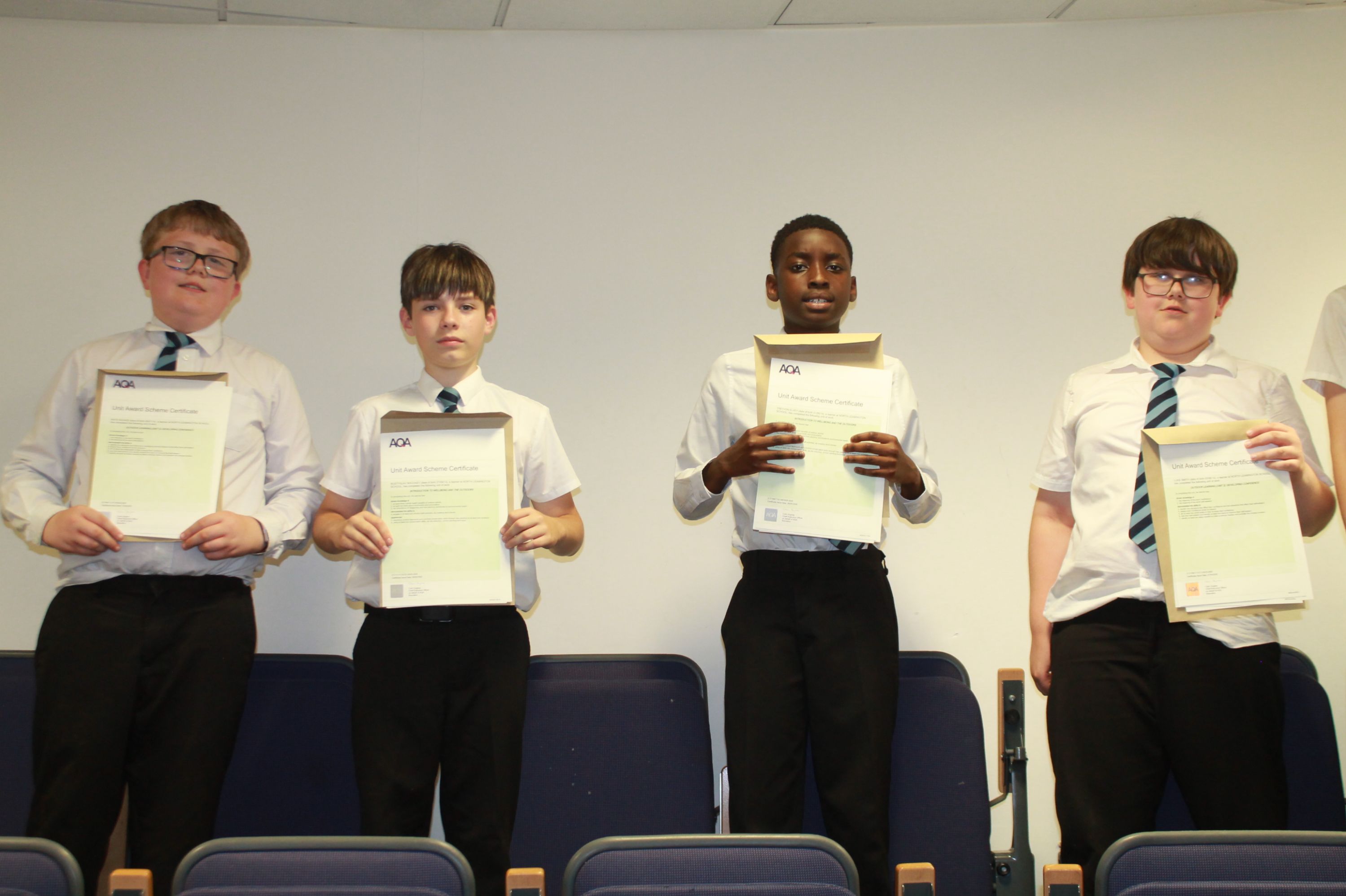Photograph of four boys standing in a row holding certificates, likely celebrating an academic or extracurricular achievement. They wear white shirts and black trousers, with a plain background and chairs partially visible behind them