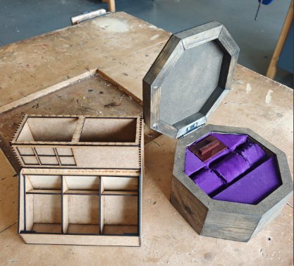 Photograph of two wooden storage boxes placed on a workbench, one rectangular with multiple compartments and the other octagonal with a hinged lid and purple fabric lining. The octagonal box contains a small wooden item, highlighting craftsmanship and organisation features of both boxes.