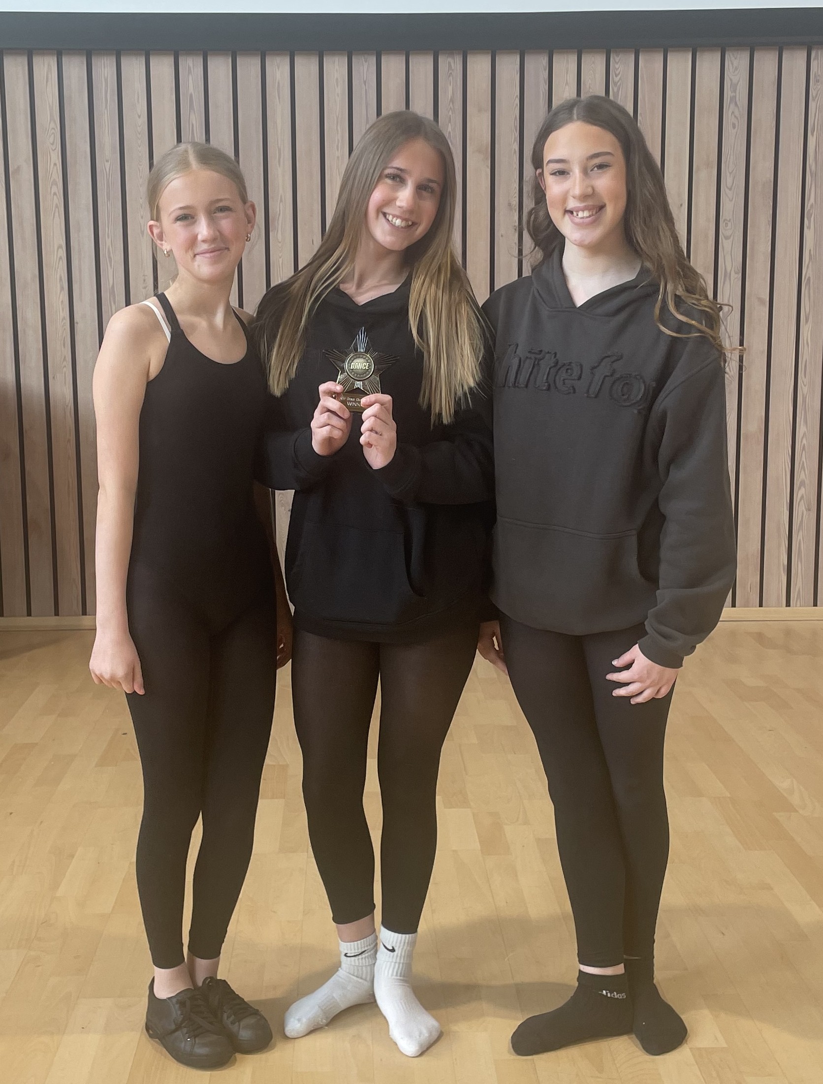 Photograph of three students standing on wooden floor in front of wooden panel wall and white screen, dressed in black dance clothing. Two wear black socks, one wears white socks, and one holds a trophy in her hand.