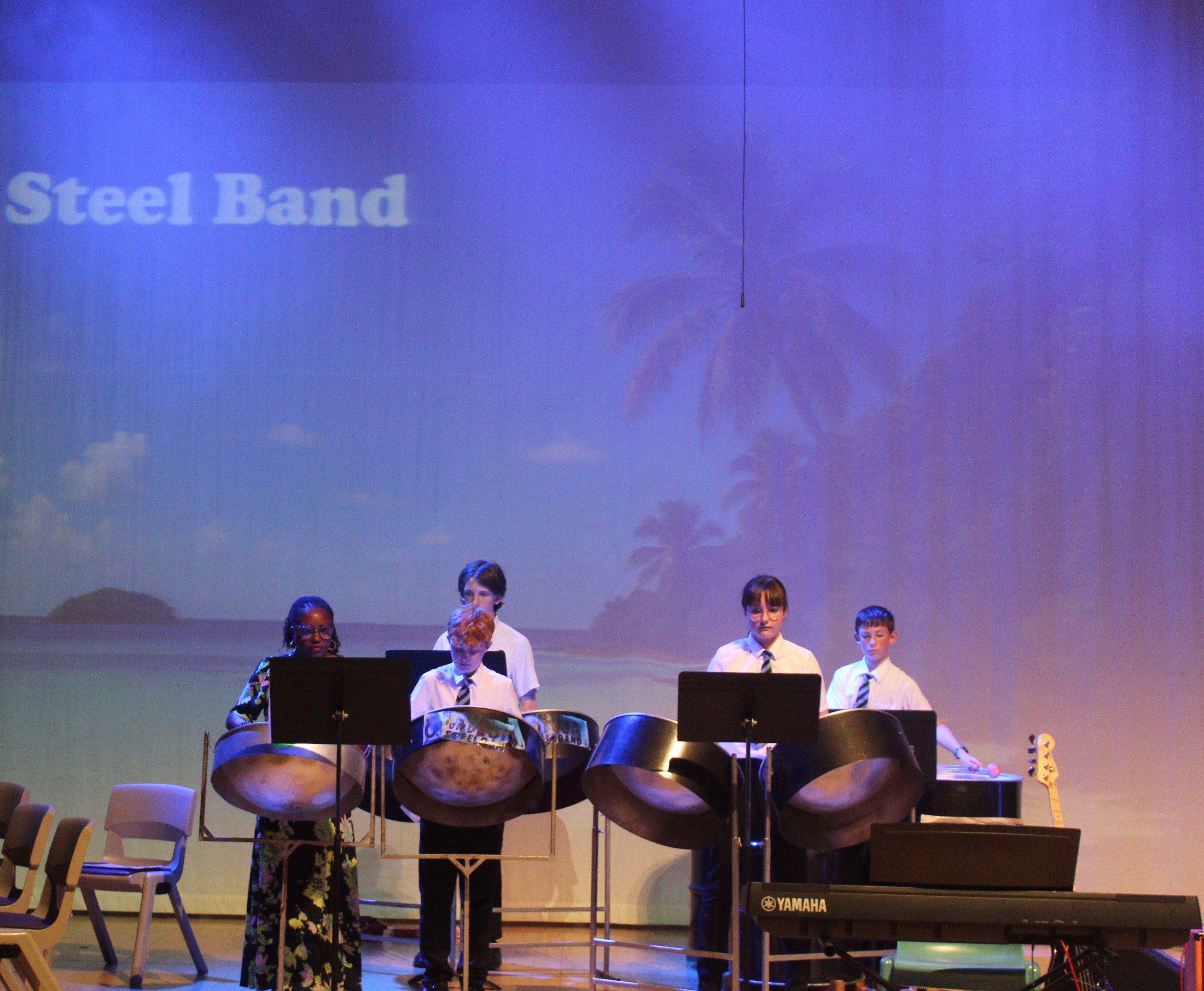 Photograph of a steel band performance on stage with four musicians playing steel pans and one playing keyboard, set against a backdrop projecting a tropical beach scene with palm trees and ocean. Text "Steel Band" appears in upper left, with musicians dressed in formal attire and music stands in front.