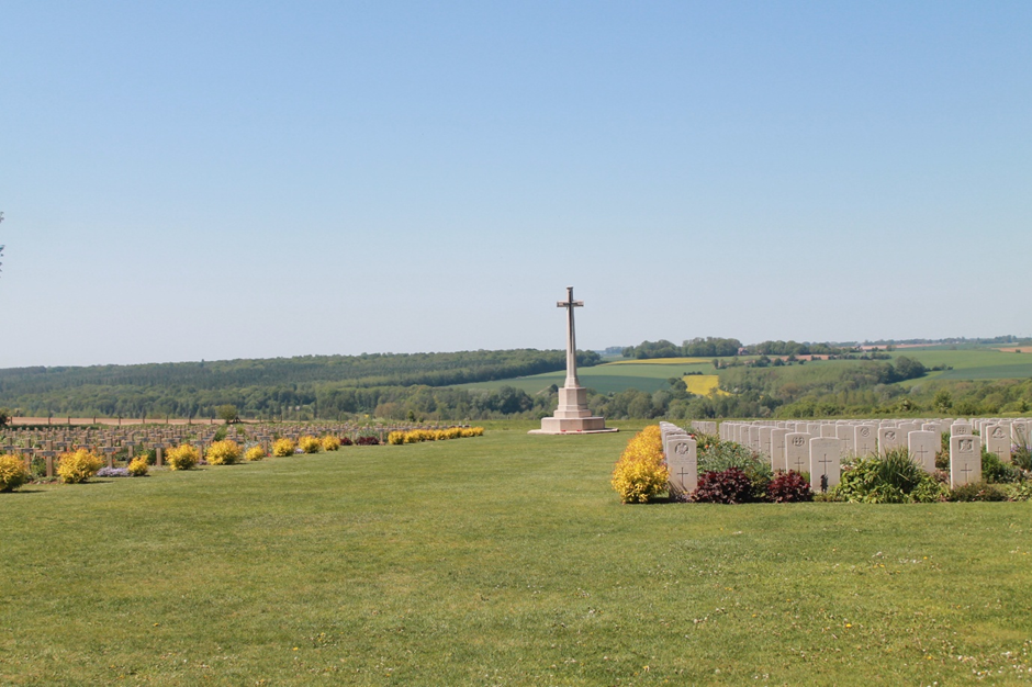 Photograph of a war cemetery featuring rows of white headstones and a tall stone cross monument in the centre. The cemetery is surrounded by green grass, yellow flowering bushes, and rolling countryside under a clear blue sky.