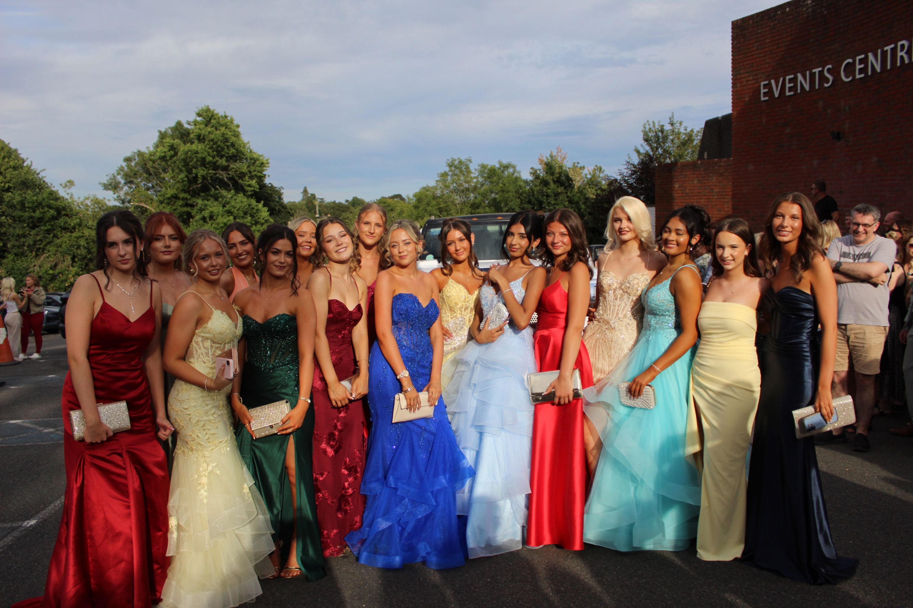 Photograph of a group of young women dressed in colourful formal gowns standing outdoors near a building labelled "Events Centre." The scene captures a celebratory moment, likely a prom or formal event, with dresses in red, yellow, green, blue, white, turquoise, and black, and the women holding small clutches.
