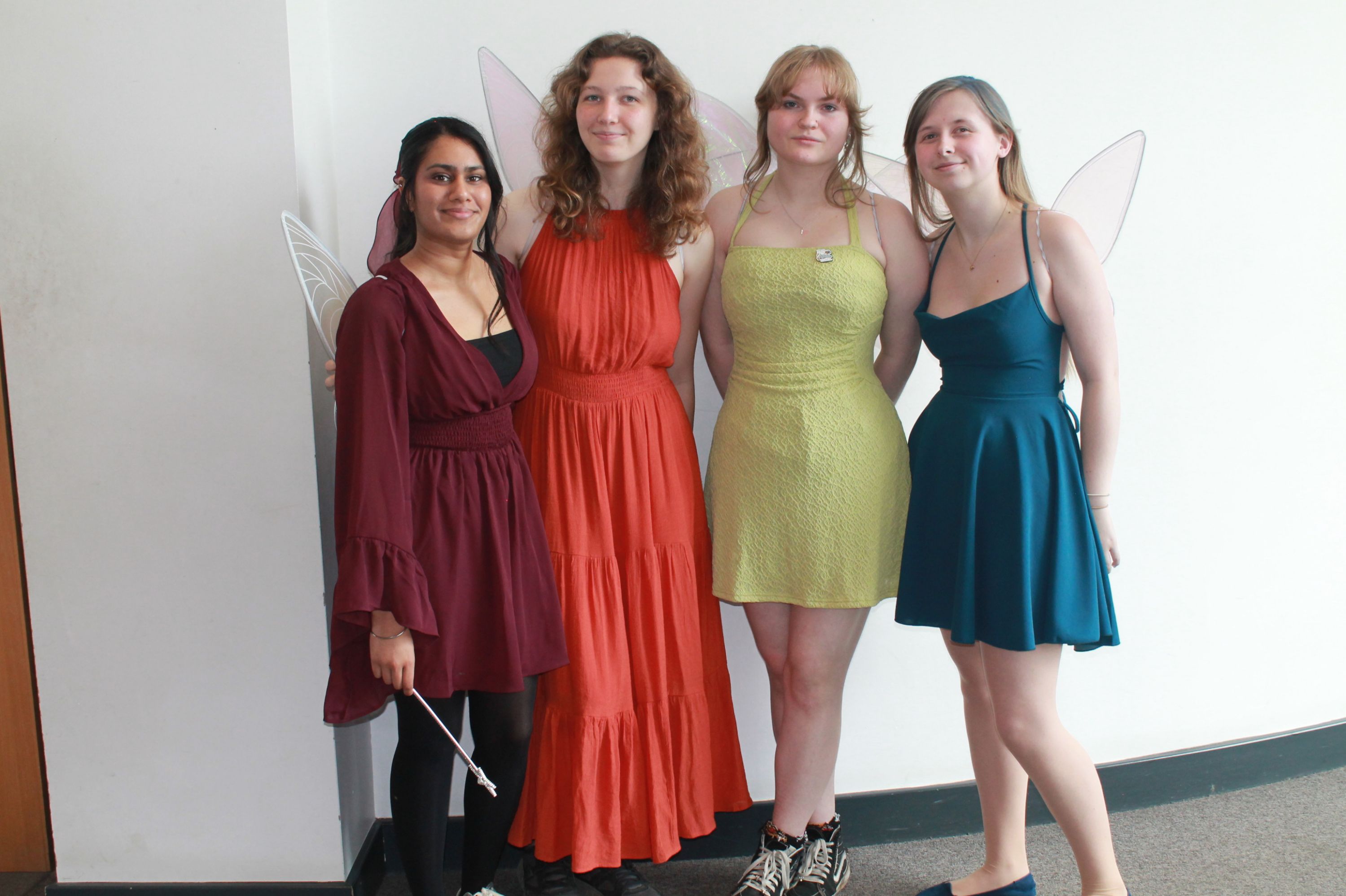 Photograph of four women standing side by side against a plain white wall, each wearing fairy wings and dresses in different colours: burgundy, orange, lime green, and teal. 
