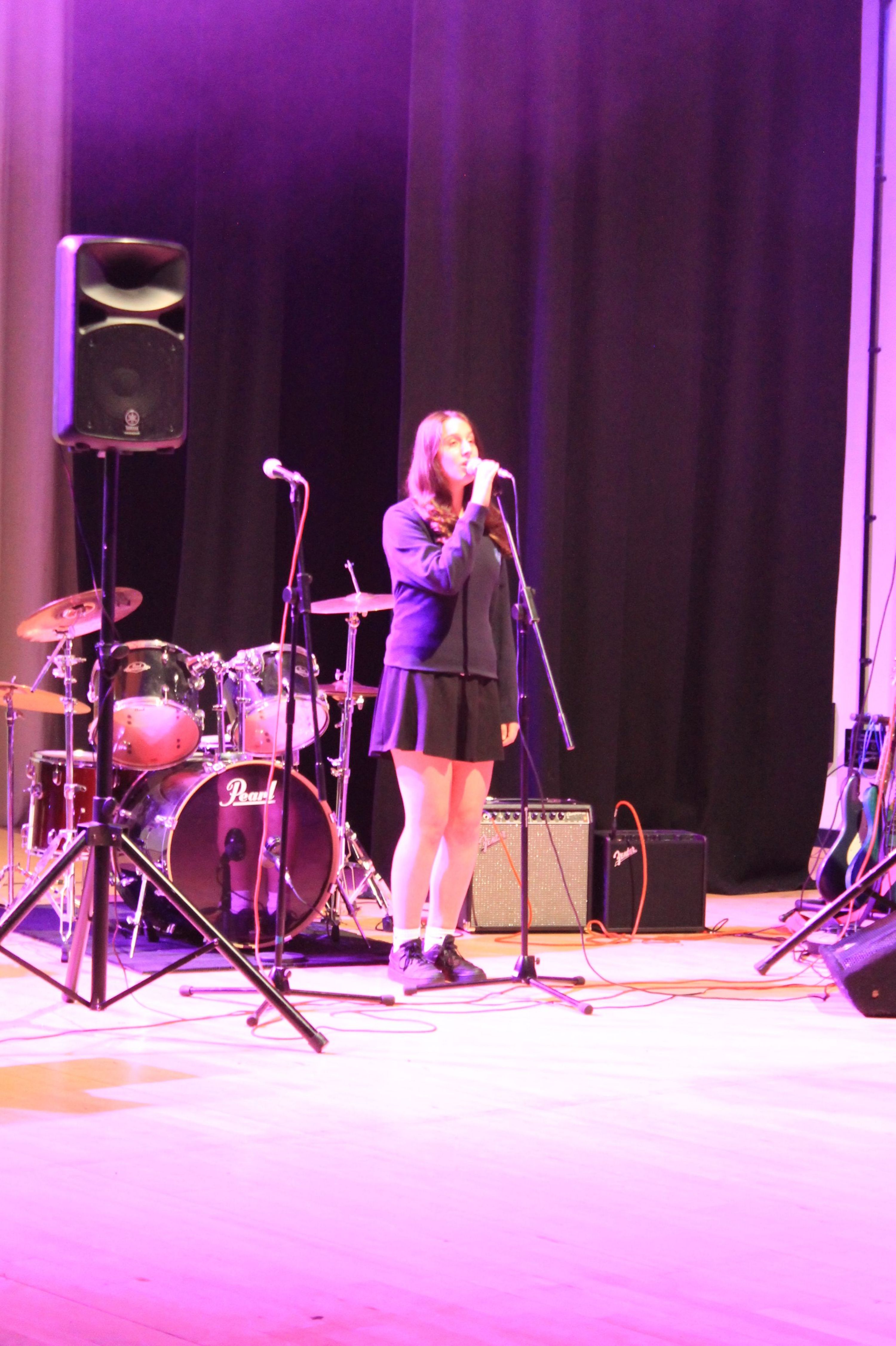 Photograph of a female performer standing on stage with a microphone, surrounded by musical instruments including a drum set and amplifiers. Stage lighting casts a purple hue, highlighting the performer dressed in a dark outfit and white shoes, suggesting a live music or vocal performance event.