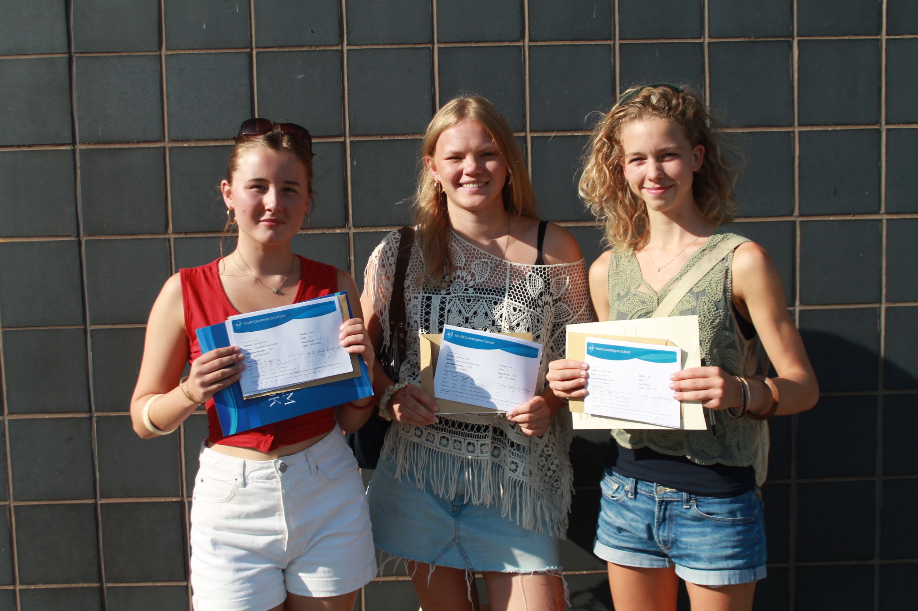 Photograph of three young women standing against dark tiled wall, each holding certificates and smiling at camera celebrating their A level result