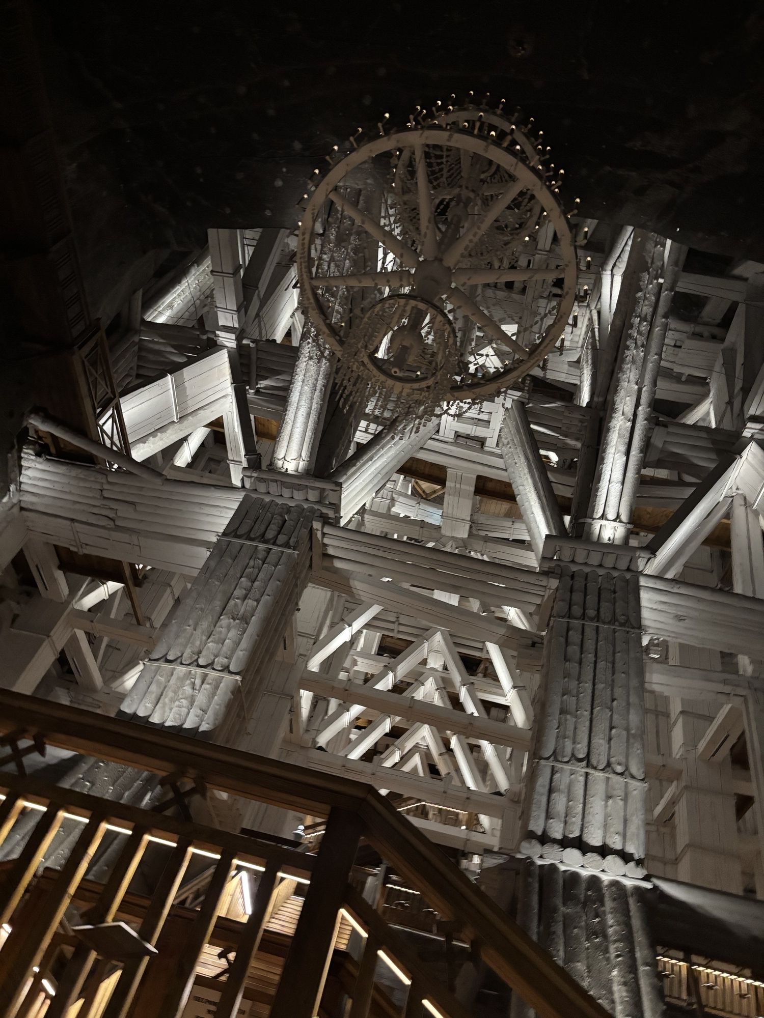 Photograph of an intricate wooden interior structure showcasing large beams, columns, and a circular wooden chandelier. The composition highlights geometric patterns formed by intersecting beams and staircases, with contrasting light and shadow emphasizing architectural details
