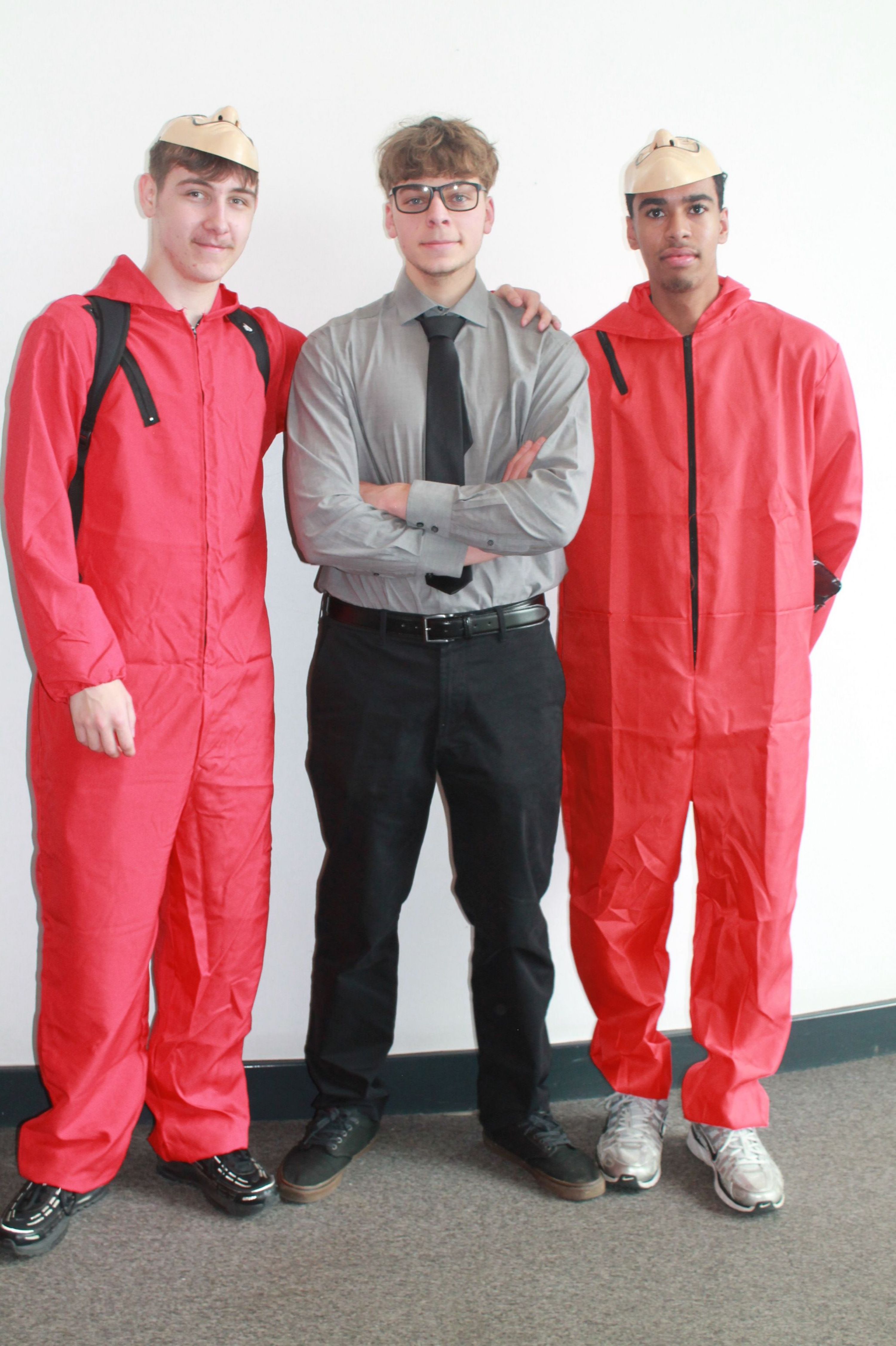 Photograph of three men standing indoors against a white wall and grey carpeted floor. Two men wear red overalls and beige caps, while the man in the centre wears a grey shirt, black tie, and black trousers.