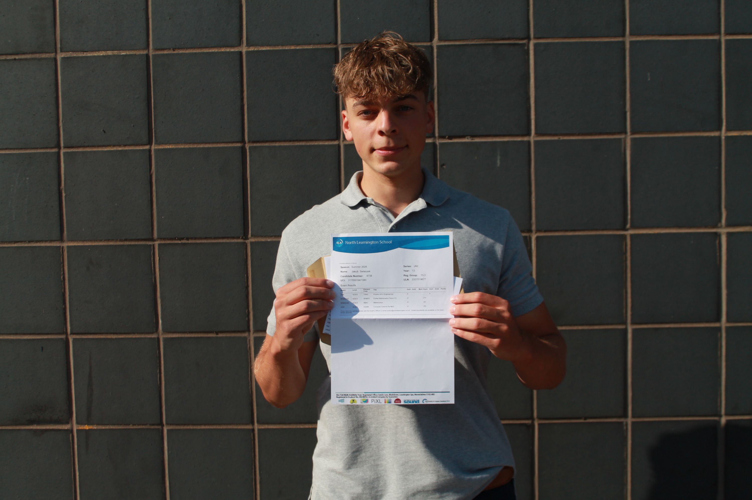 Photograph of a young man standing against a dark tiled wall holding a document with both hands