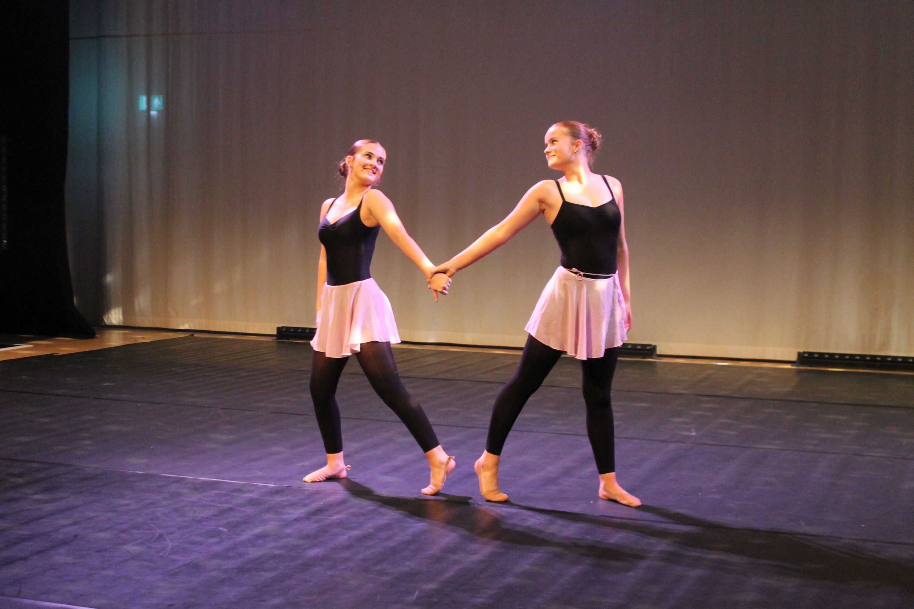 Photograph of two dancers performing on stage, wearing black leotards, pink skirts, and black leggings. They hold hands while extending opposite legs and looking away from each other, with a plain curtain backdrop and stage lighting
