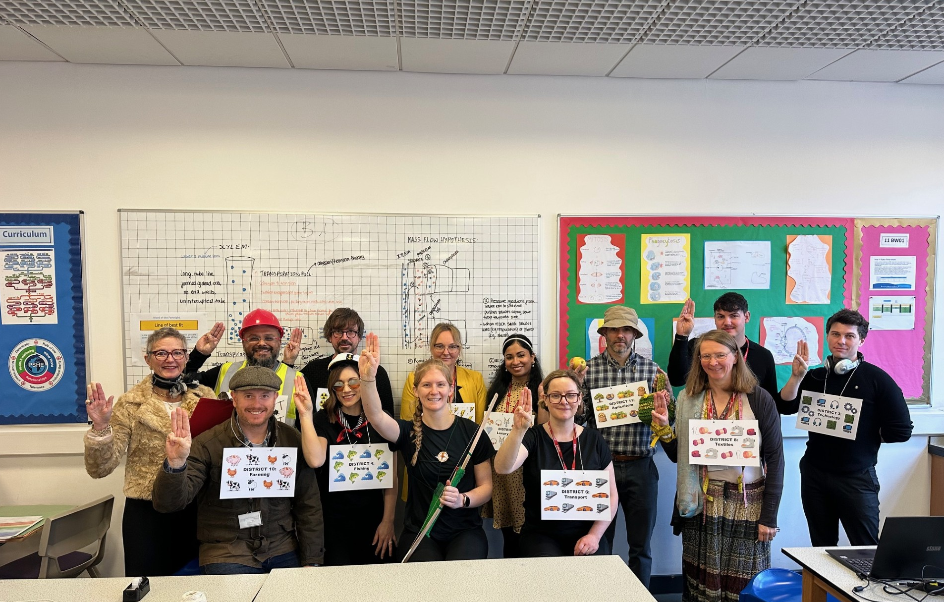 Photograph of a group of people in a classroom setting holding signs with various symbols and illustrations, some raising three fingers. Classroom walls display charts, graphs, and colourful bulletin boards with educational content, indicating a learning or awareness activity.