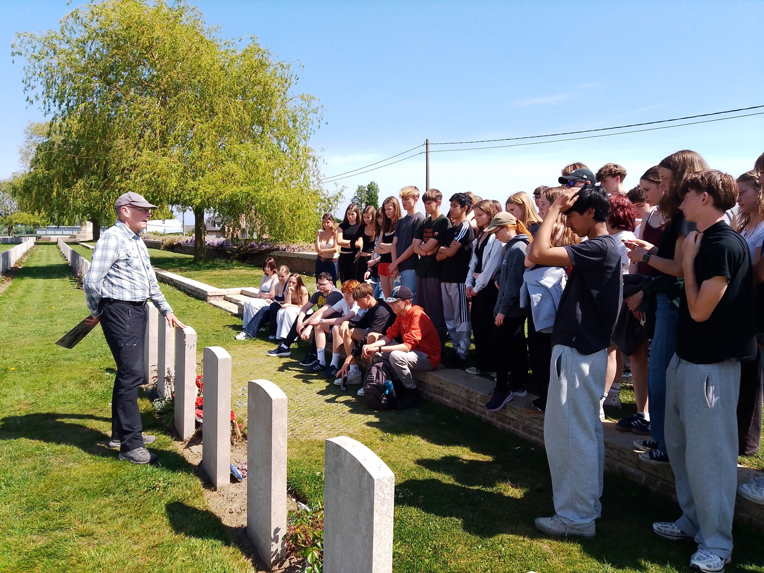 Photograph showing a group of young people gathered outdoors at Lijssenthoek Military Cemetery listening to an older man speaking near a row of gravestones, suggesting an educational or commemorative event. The setting includes green grass, a large tree, and clear blue sky, with participants standing and sitting attentively along a stone ledge.