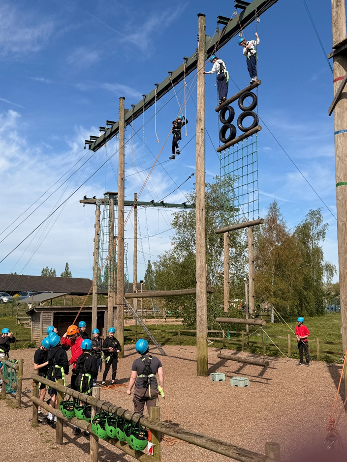 Photograph of an outdoor ropes course showing several participants wearing helmets and harnesses engaged in climbing and balancing activities on wooden poles, nets, and suspended tires. Bright blue sky and green trees form background, with instructors or supervisors visible on the ground ensuring safety.