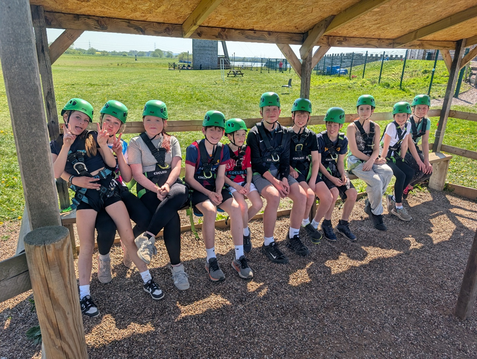 photograph showing a group of children wearing green helmets and harnesses sitting on a wooden bench under a shelter, likely preparing for an outdoor adventure or climbing activity. Bright sunny day with grassy field and climbing structures visible in background, highlighting a recreational or team-building setting.