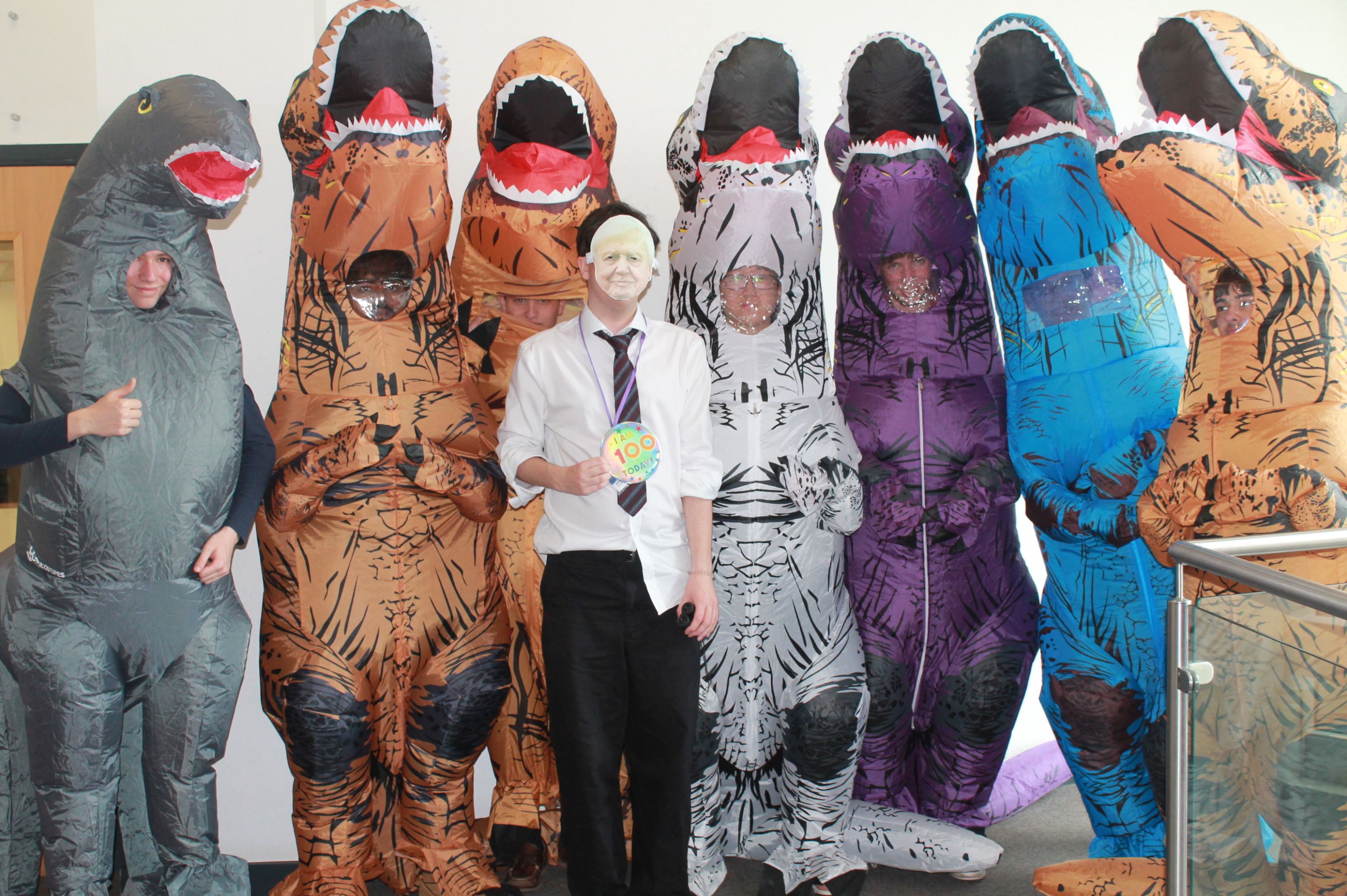Photograph showing seven people dressed in inflatable dinosaur costumes in various colours including grey, brown, white, purple, and blue, standing next to a person in formal attire holding a "100" badge
