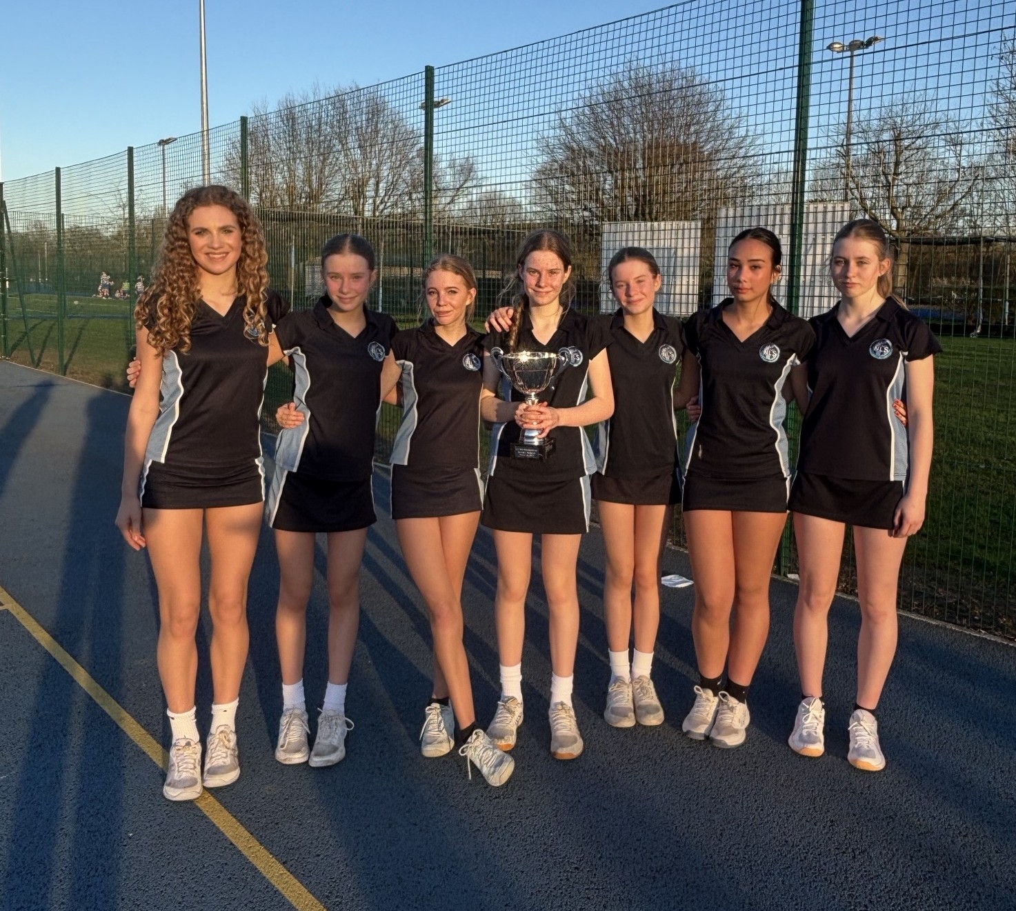 Photograph of seven netball players standing in a row on an outdoor court, wearing matching black uniforms with white sneakers. One player in the centre holds a trophy, with a clear blue sky and netted fence in background.