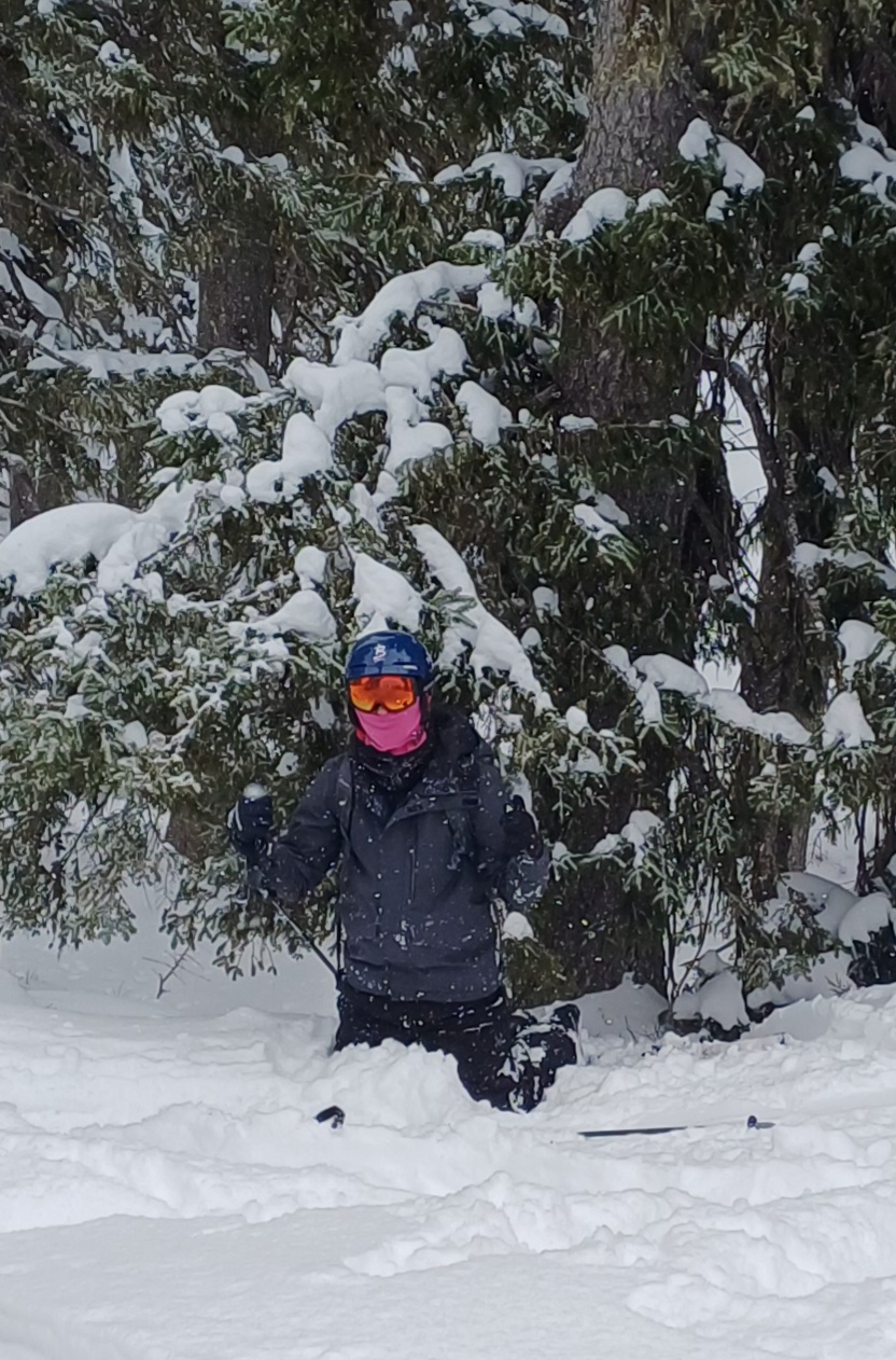 Photograph of a person skiing in deep snow surrounded by snow-covered evergreen trees. The skier wears a blue helmet, orange goggles, a pink face covering, and dark winter clothing, kneeling in snow with ski poles visible.
