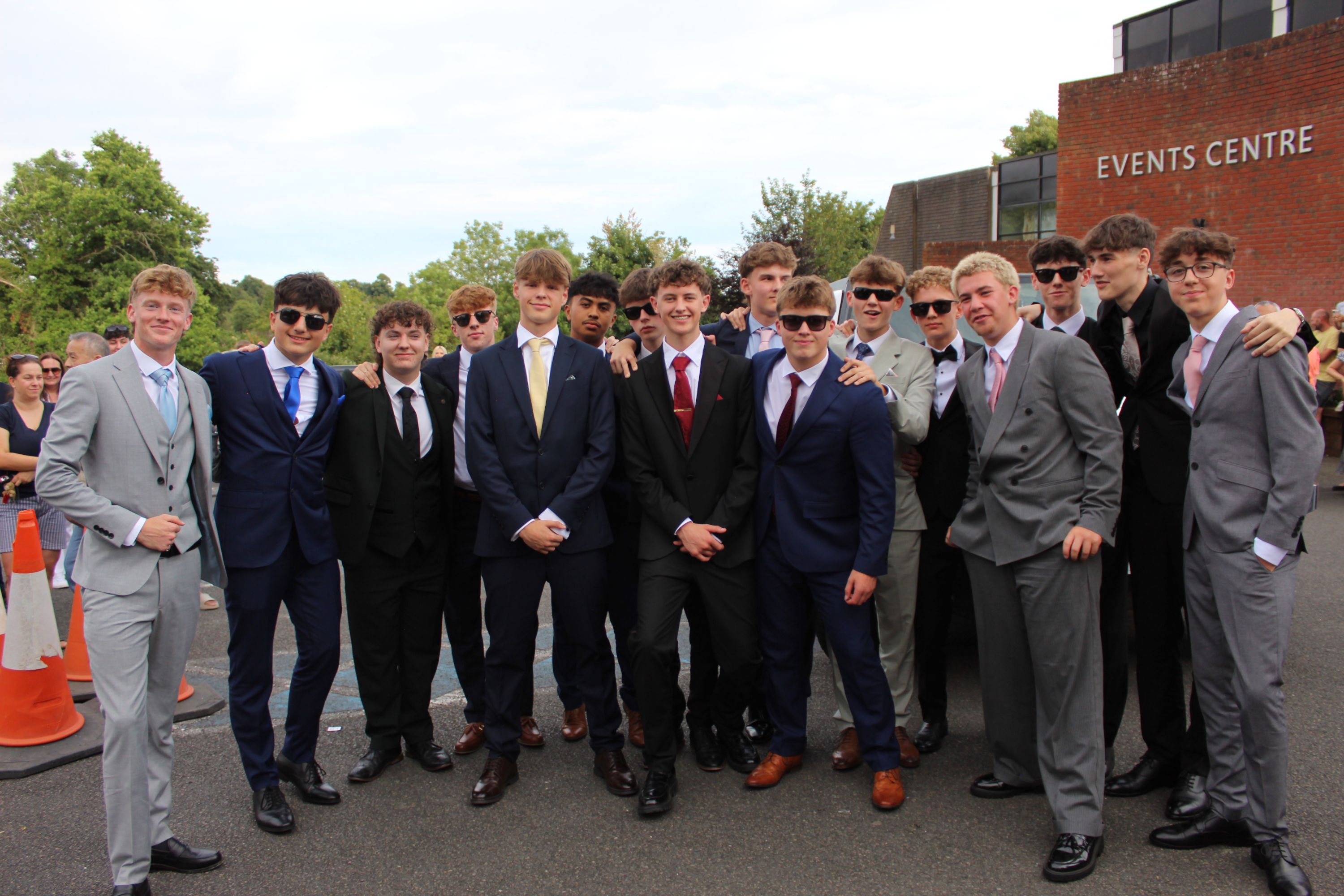 Photograph of a group of young men dressed in formal suits and sunglasses, standing outdoors near a building labelled "Events Centre." The group appears to be attending a special occasion, with a mix of dark and light suits, ties, and polished shoes, set against a backdrop of trees and traffic cones.