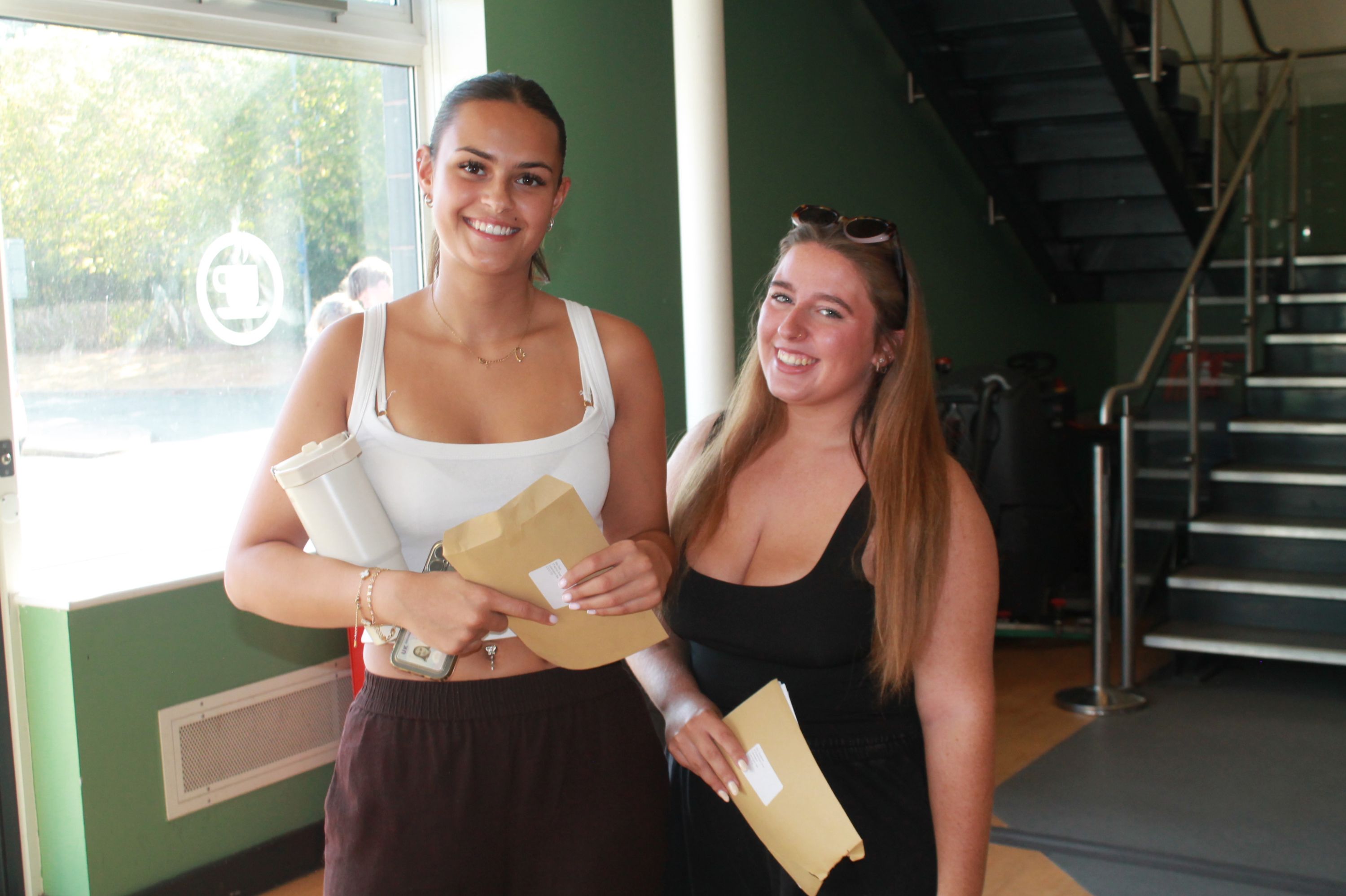 Photograph showing two young women smiling and holding A level results standing indoors near a staircase with green walls in background. Both wear casual tops, one white and one black, 