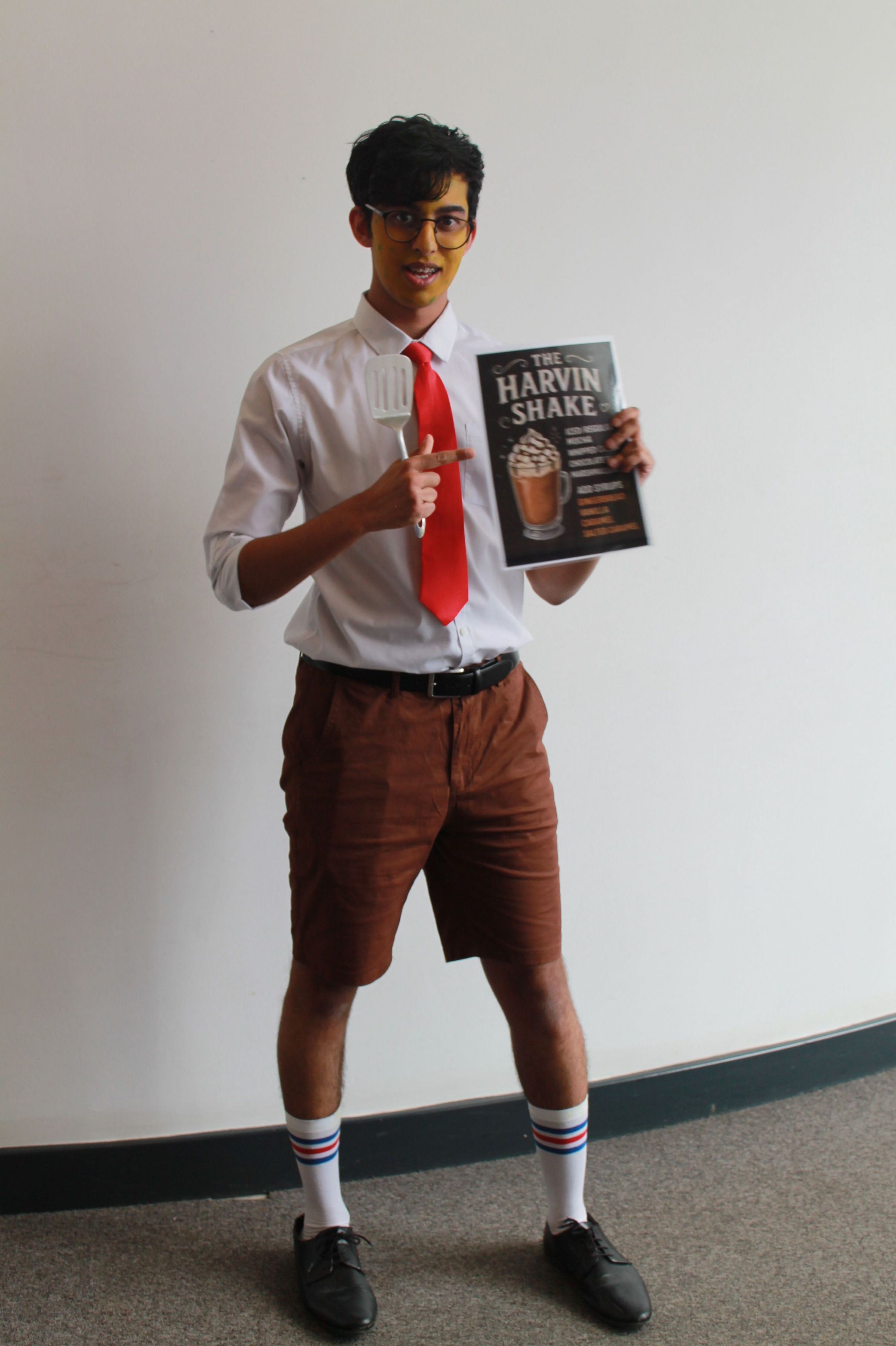 Photograph of a person dressed in a white shirt, red tie, brown shorts, and striped socks holding a book titled "How to Win Friends and Influence People." The person is standing against a plain white wall, smiling, and pointing upwards with one finger.