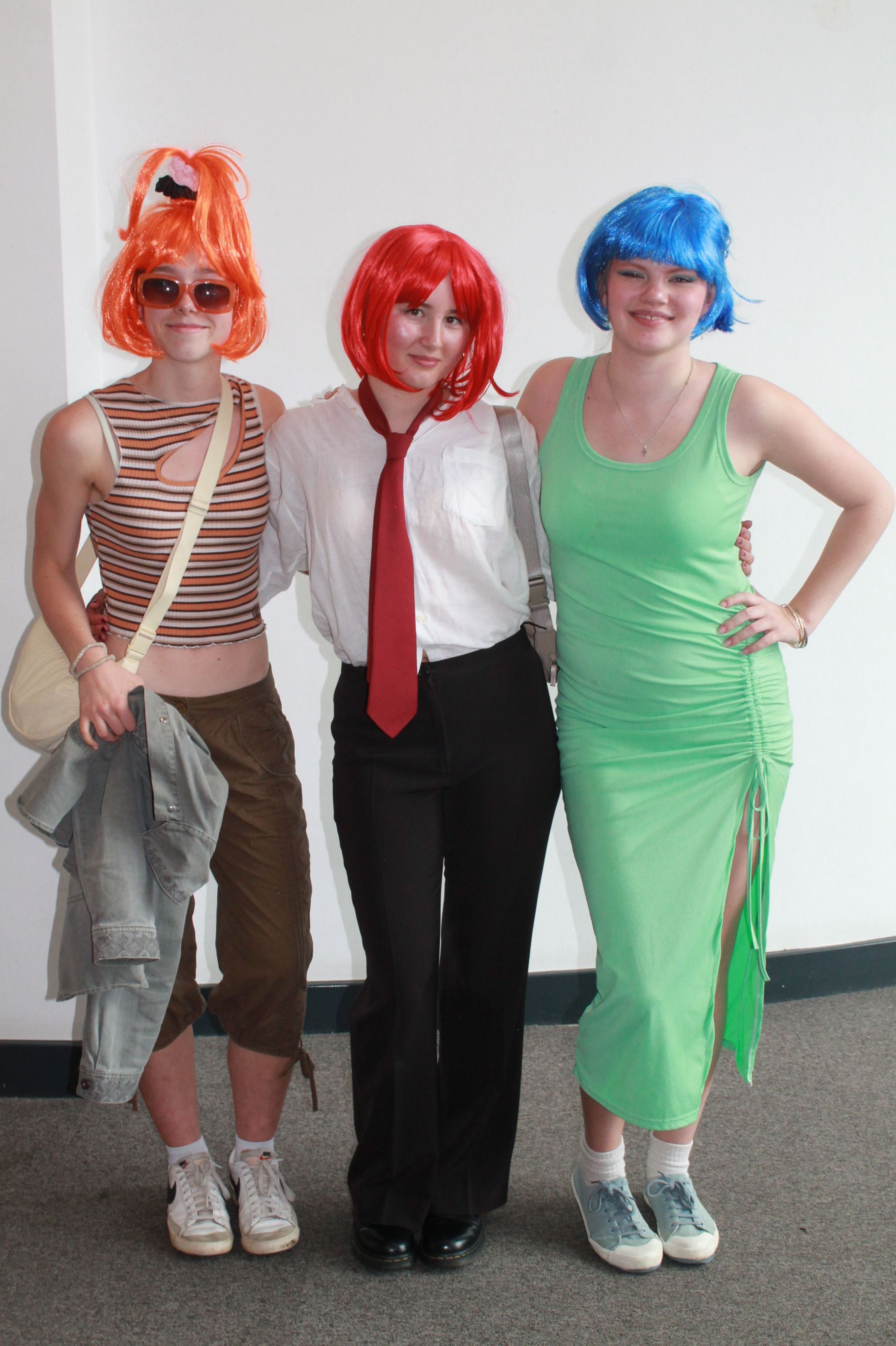 Photograph of three people standing indoors against white wall and grey carpet floor, each wearing brightly coloured wigs and casual or costume clothing, including blue wig with green dress, red wig with white shirt and red tie, and orange wig with striped top and sunglasses.