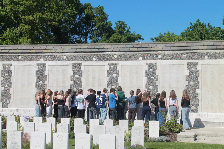 Photograph of a group of young people gathered in front of a stone memorial wall engraved with numerous names at Tyne Cot, paying respects at a war cemetery. White headstones with engraved symbols and inscriptions are visible in the foreground, set against a backdrop of green trees and clear blue sky.