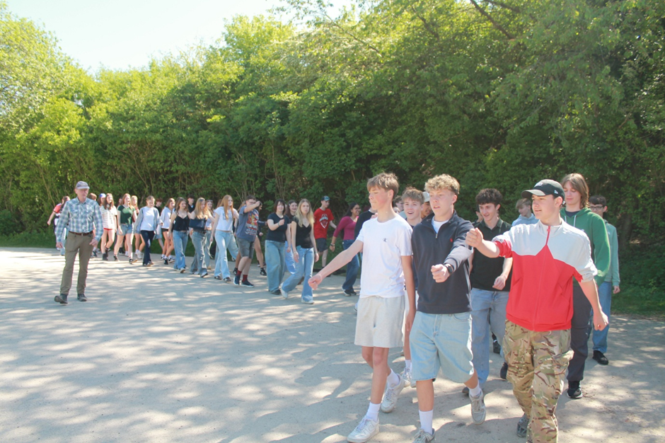 Photograph showing a large group of young people walking outdoors on a sunny day, led by three boys in casual clothing and an older man walking behind them. Background features dense green trees, and the group appears to be participating in an organised marching activity.