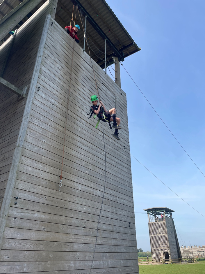 Photograph showing two people abseiling down a tall wooden climbing wall under clear blue sky. Both individuals wear helmets and harnesses, with one person descending while the other assists from the top platform. 