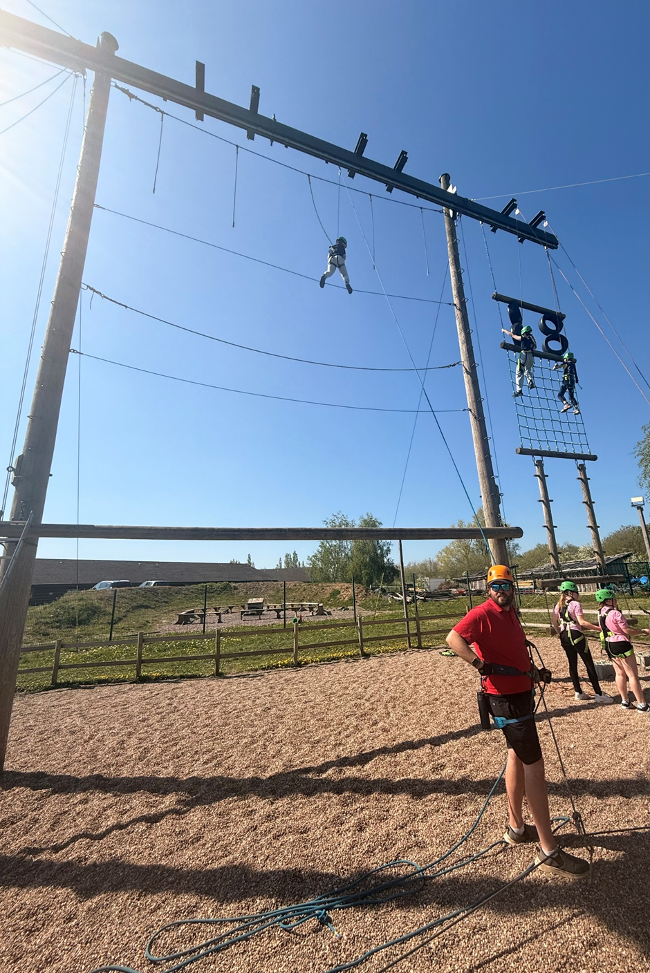 Photograph of an outdoor ropes course featuring participants climbing and suspended on various elements, including a high wire and cargo net. Bright blue sky and safety personnel wearing helmets and harnesses are visible, highlighting an active team-building or adventure activity setting.