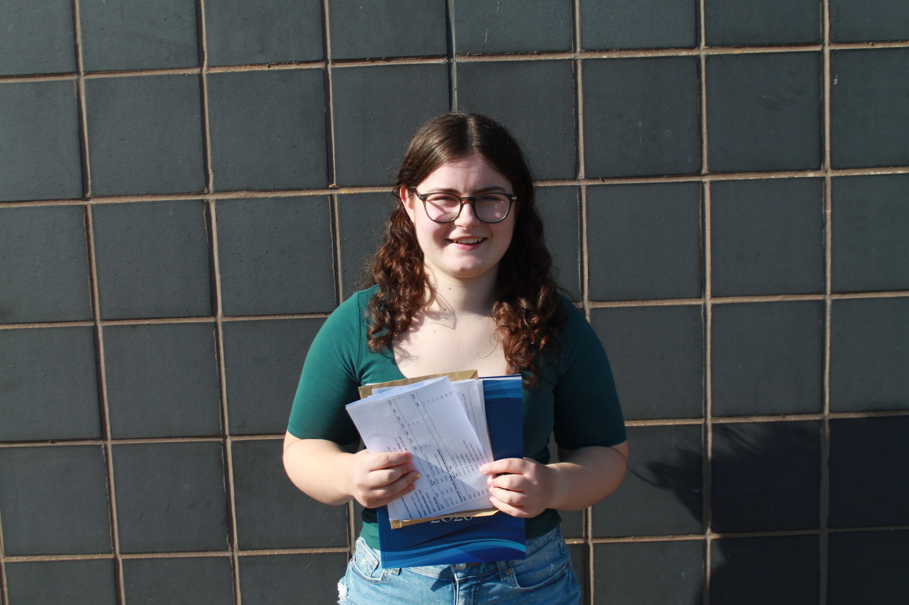 Photograph of a young woman standing against a dark tiled wall holding a document with both hands