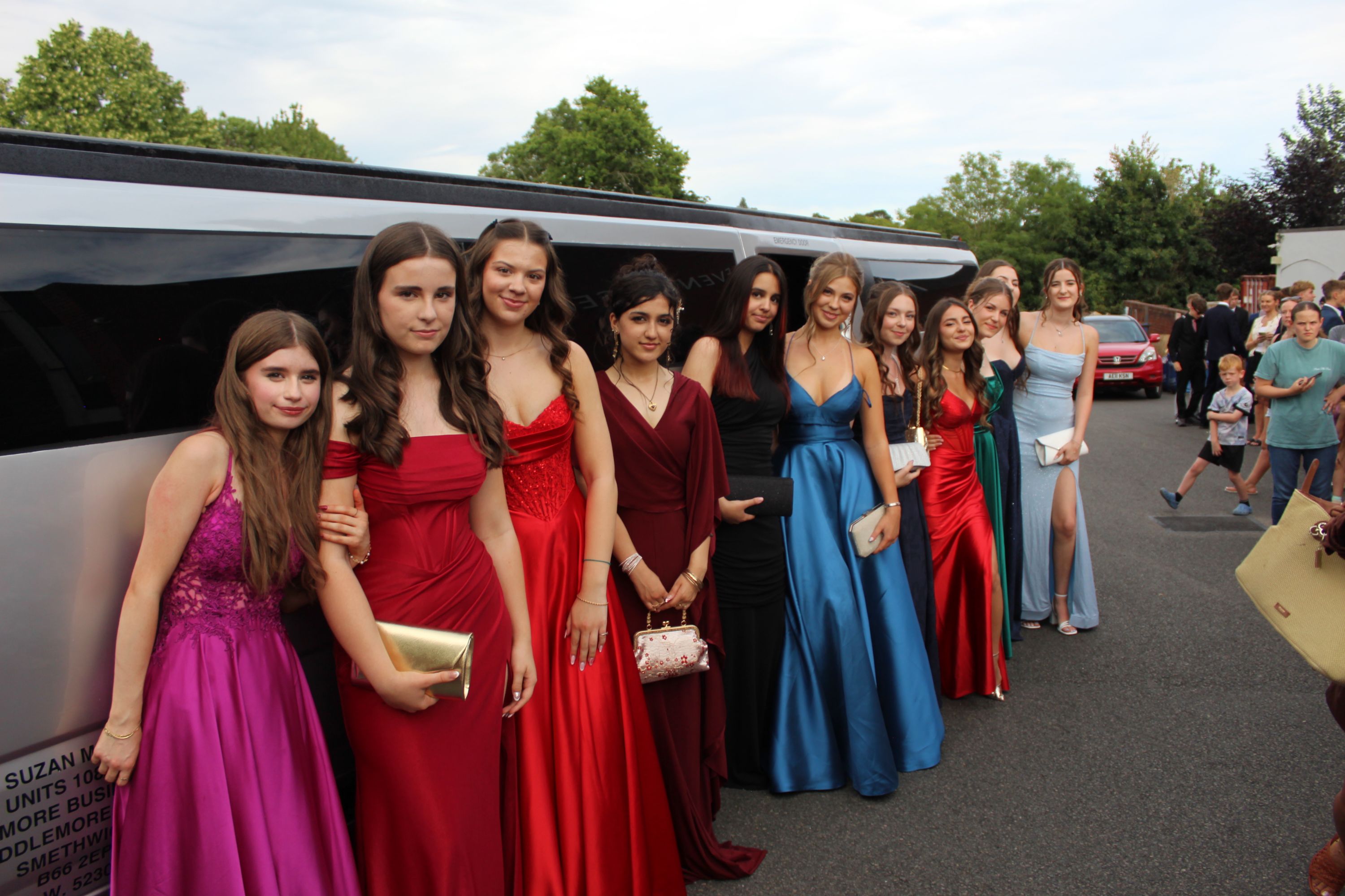 Photograph of a group of young women dressed in formal gowns standing in a line beside a limousine outdoors. Dresses feature various colours including purple, red, blue, and light blue, with some holding clutches, at prom 