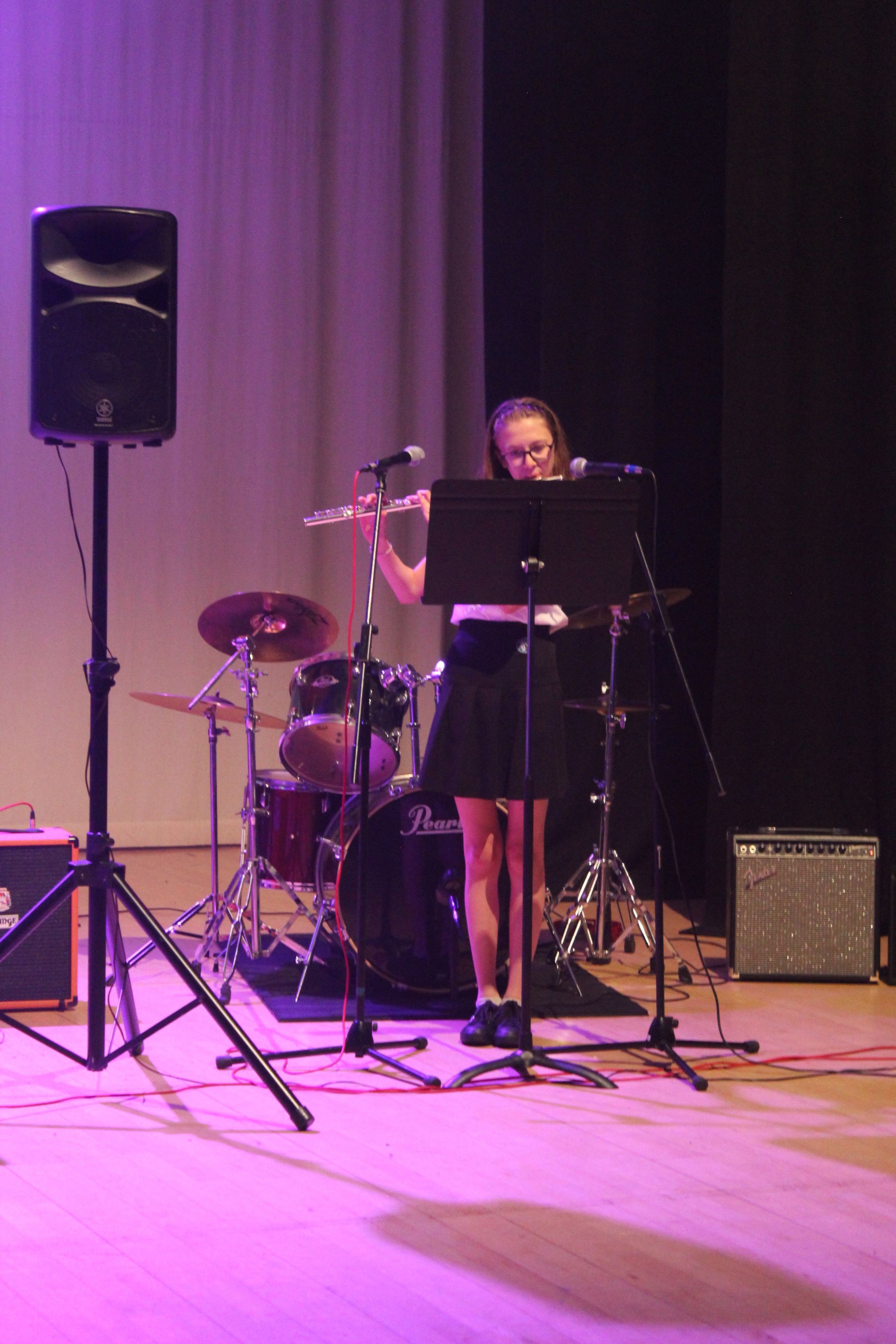 Photograph of a young girl playing a flute on stage during a musical performance, accompanied by a drum set and amplifier in the background. Stage lighting casts a purple hue on wooden floor, with microphone stands and speaker visible, highlighting a live concert setting.