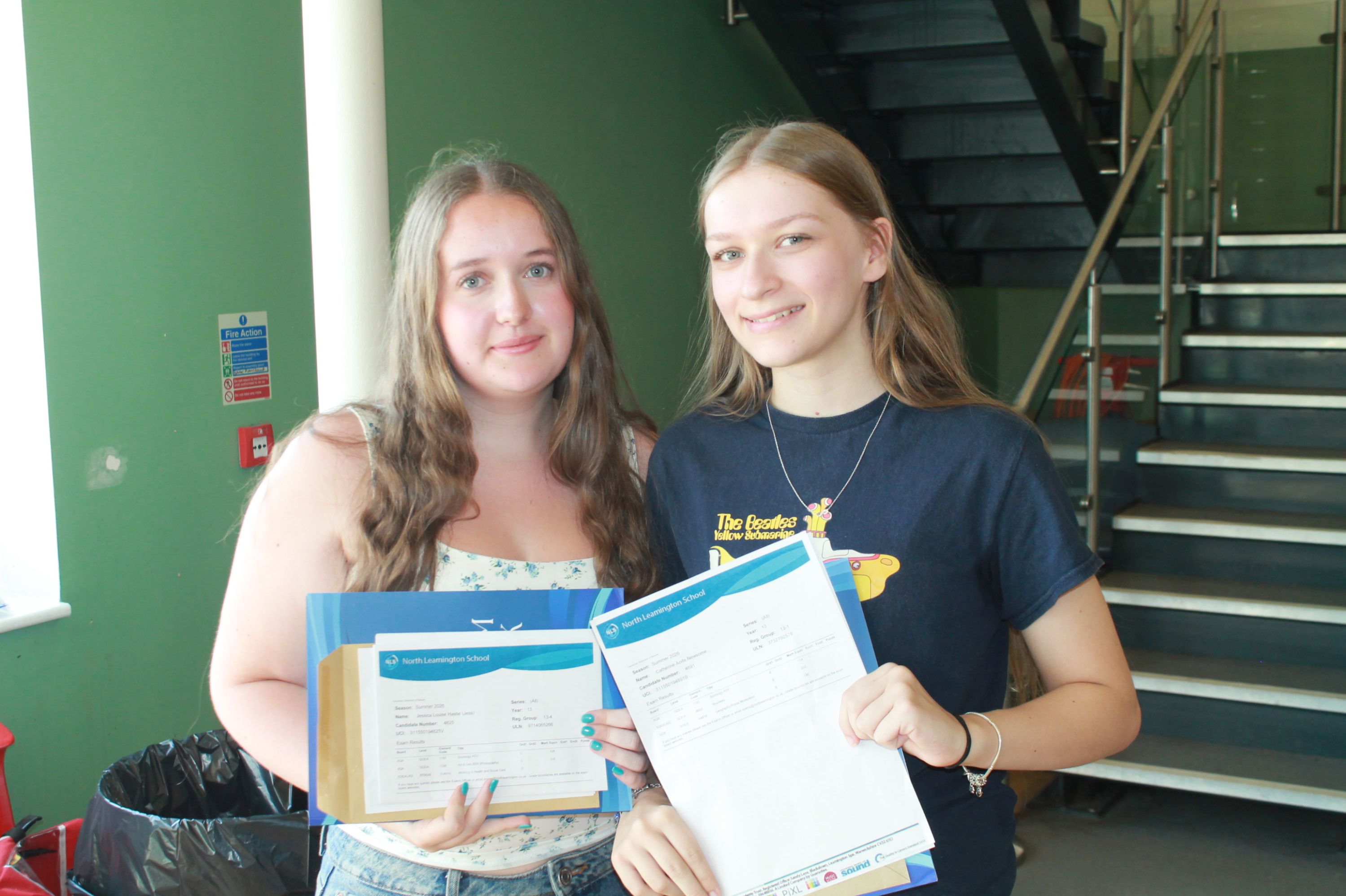 Photograph showing two young women smiling and holding A level results standing indoors near a staircase with green walls in background. Both wear casual tops, one white and one black, 