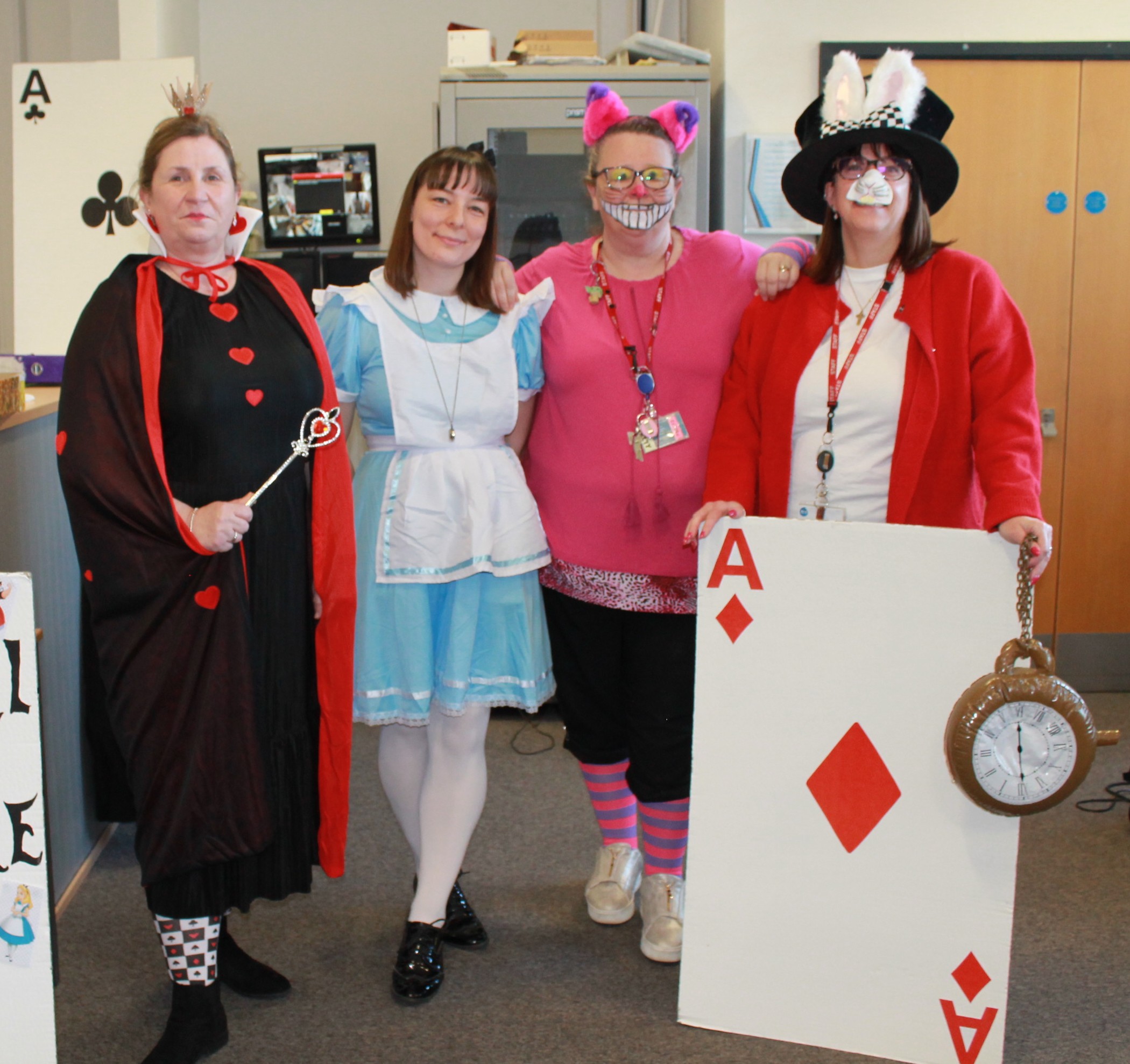 Photograph of four people dressed in Alice in Wonderland-themed costumes standing in an office setting. Costumes include Queen of Hearts, Alice, Cheshire Cat, and Mad Hatter holding a large Ace of Diamonds card and a pocket watch, with a sign reading "We're All Mad Here" featuring characters from the story.