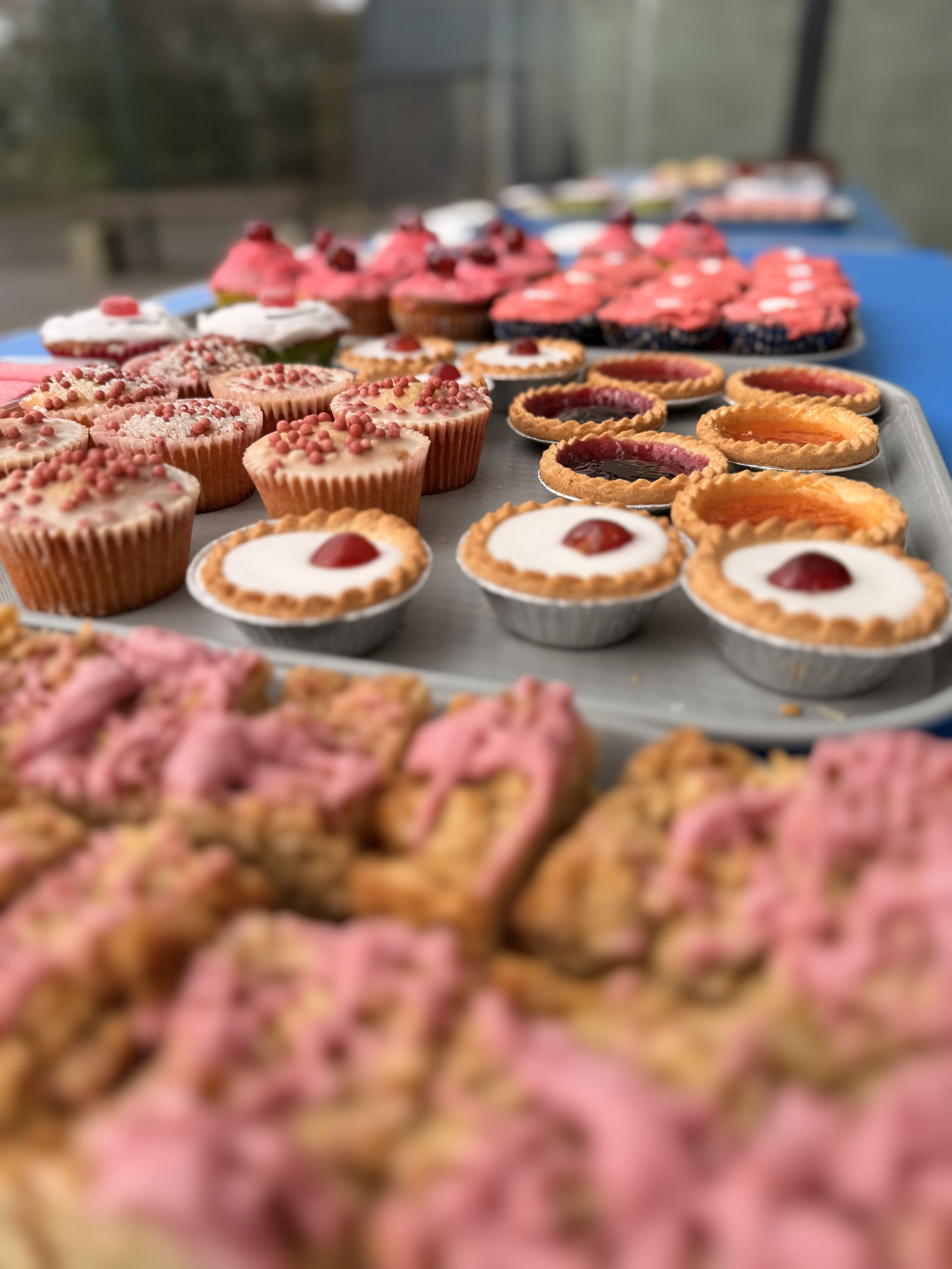 Photograph of assorted baked goods arranged on trays, showcasing cupcakes, tarts, and pastries with various toppings and icing. Focus highlights pink and red decorations on cupcakes and tarts, with blurred pastries in foreground and background,