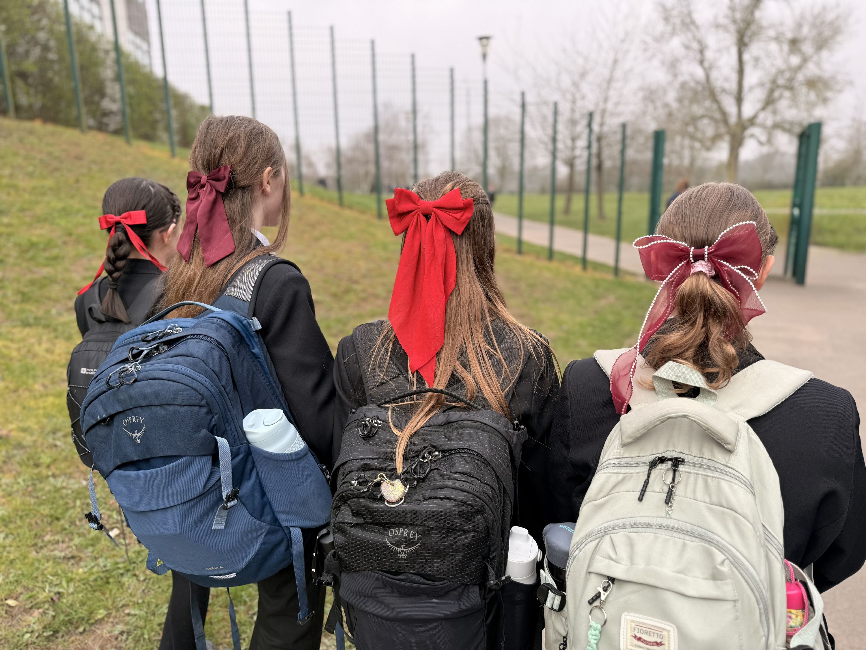 ​​​​Photograph of four students outdoors on a path beside a fenced grassy area, each wearing a school uniform and carrying backpacks in blue, black, and white. All girls have large red bows tied in their hair.
