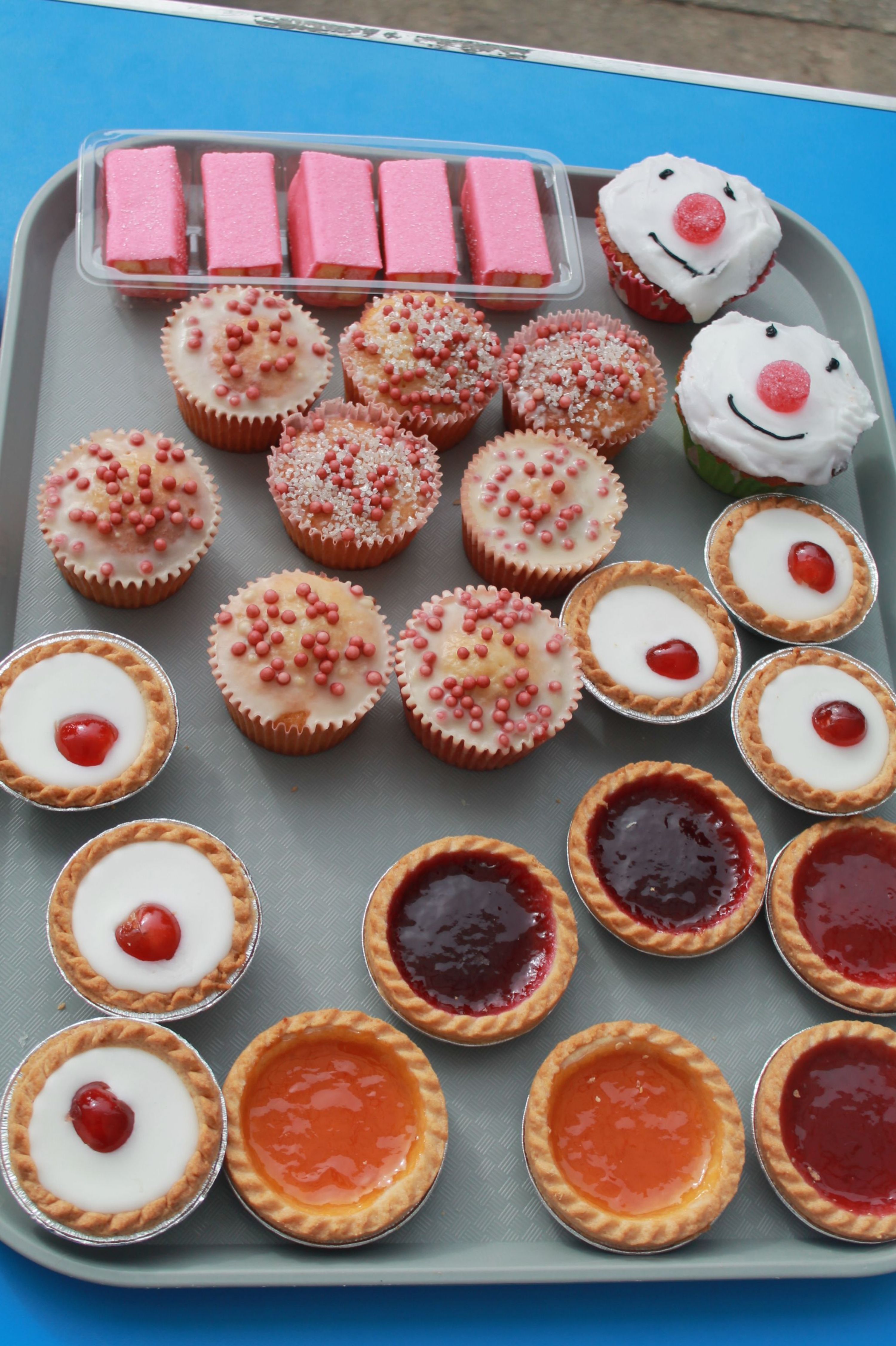 Photograph of a tray filled with assorted baked goods including cupcakes, jam tarts, and pink wafer biscuits arranged in neat rows. Cupcakes feature various decorations such as white icing with red sprinkles and smiley faces, while jam tarts display different fruit fillings like strawberry, raspberry, and apricot.
