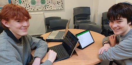 Photograph showing two people seated at a round wooden table, each using a laptop and a tablet for work or study. The setting appears to be a casual office or classroom with chairs and a poster on the wall in the background.