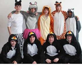 Photograph of a group of ten people dressed in various animal costumes, posing indoors against a plain wall and door. Costumes include zebra, mouse, lion, giraffe, penguin, and others, with five individuals standing and five kneeling,
