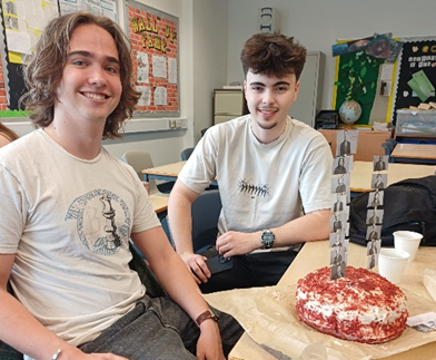 Photograph showing two young men sitting at a classroom table with a red velvet cake decorated with a tall vertical photo strip of a person in various poses. Classroom background includes bulletin boards, a globe, and chairs