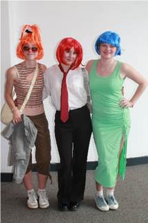 Photograph of three people dressed in colourful wigs and costumes, posing indoors against a plain white wall and grey carpet. Notable details include a blue wig with a green dress, a red wig with a white shirt and red tie, and an orange wig with sunglasses, striped top, and brown shorts.