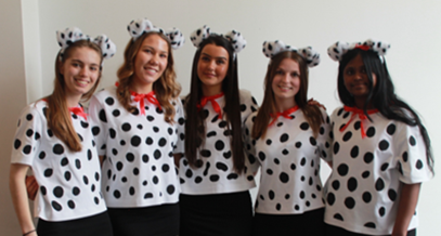 Photograph of five people standing side by side wearing matching white tops with large black polka dots and red bows, along with headbands featuring black and white spotted ears. 